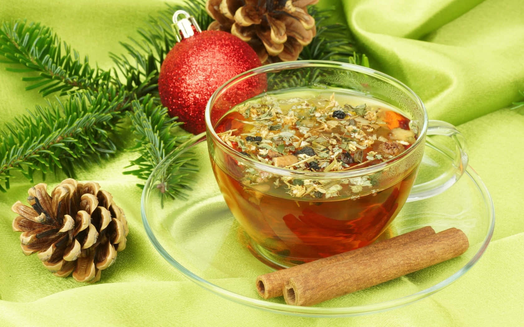Mulled Wine 桌布 壁紙 — 一杯透明玻璃杯裝著的香料草本茶，帶有漂浮的植物成分，放在碟子上，旁邊有肉桂棒、松果和一個紅色裝飾品，置於柔軟的綠色布料上。