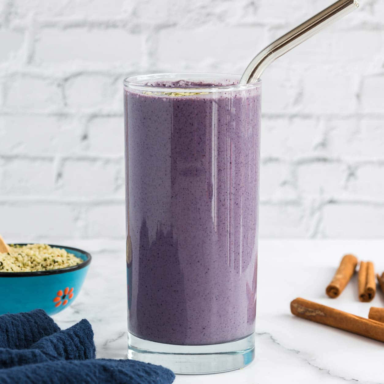 Blueberry Smoothie 桌布 壁紙 — 一杯高身嘅紫色冰沙，用金屬飲管，放喺白色雲石枱面，旁邊有肉桂棒同一個藍色碗裝住種子。