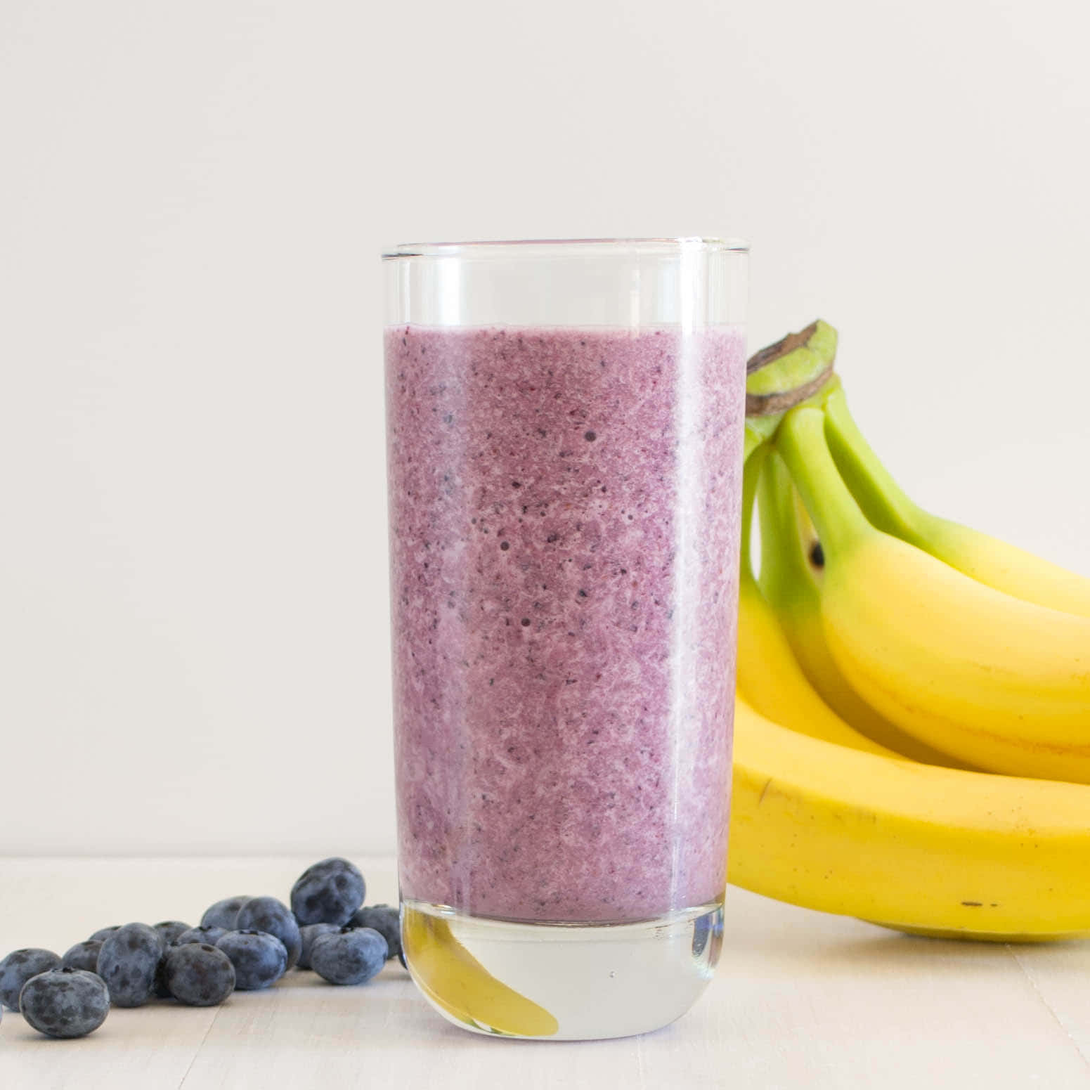 Blueberry Smoothie 桌布 壁紙 — 一杯高身的紫色藍莓香蕉冰沙，旁邊放著新鮮藍莓和一束成熟的黃色香蕉，置於淺色木質表面上。