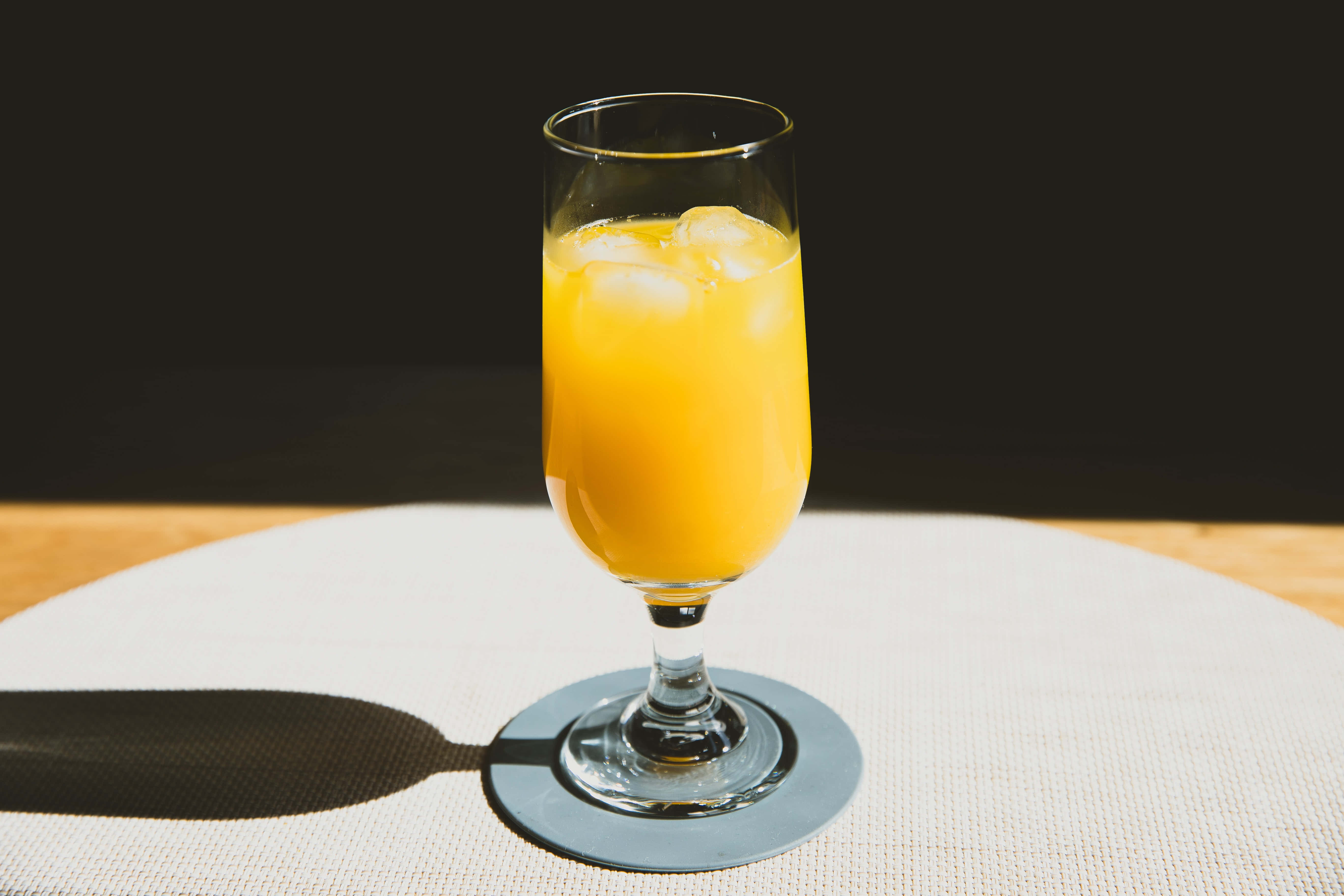 Summer Drinks 桌布 壁紙 — 一杯高身鮮橙汁加冰塊放在杯墊上，在深色背景下，於有紋理的白色餐墊上投下清晰的陰影。