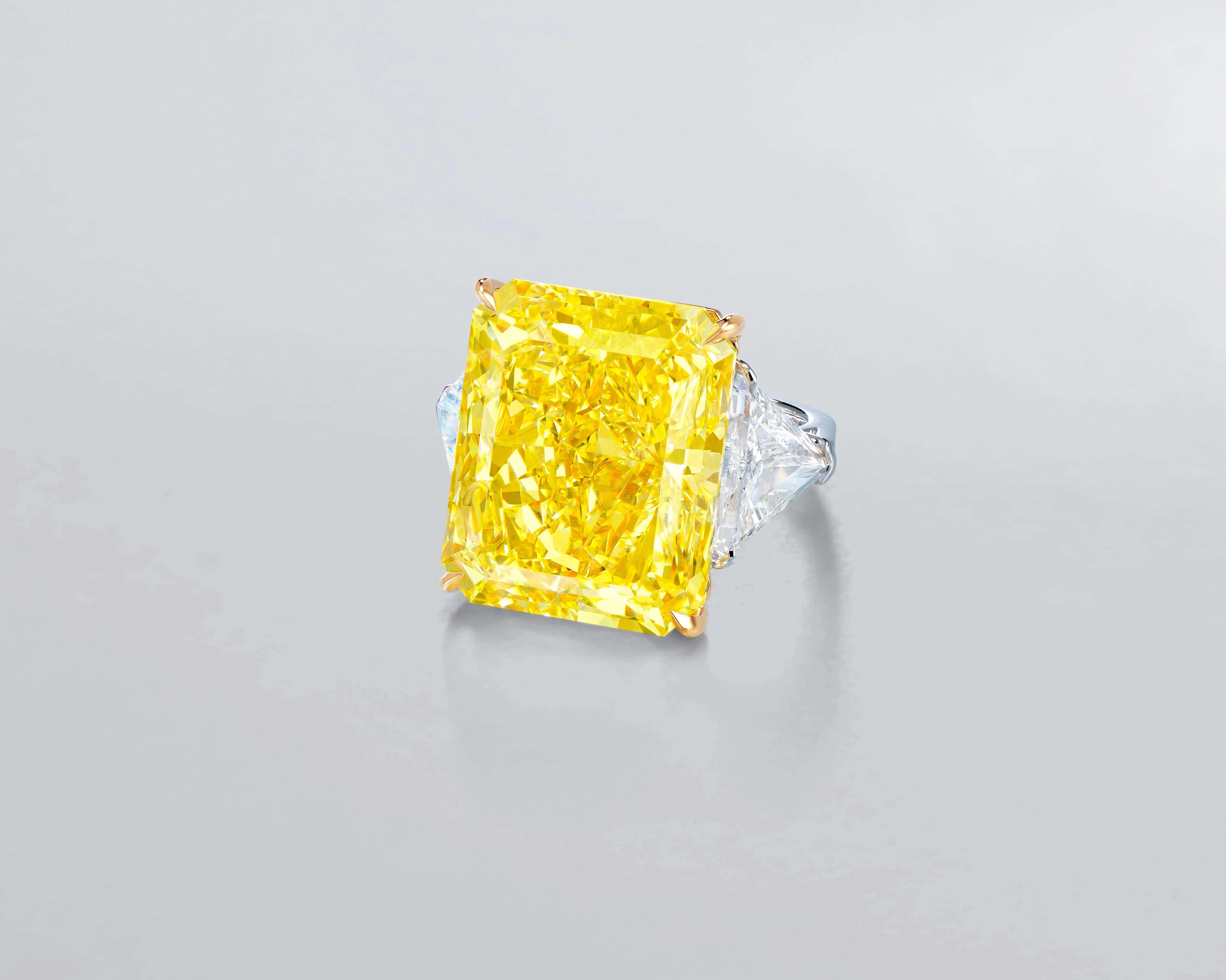 Yellow Diamond 桌布 壁紙 — 一枚令人驚豔的黃色祖母綠切割鑽石戒指，搭配兩顆清澈的三角形側石，在柔和的灰色背景上閃耀光芒。