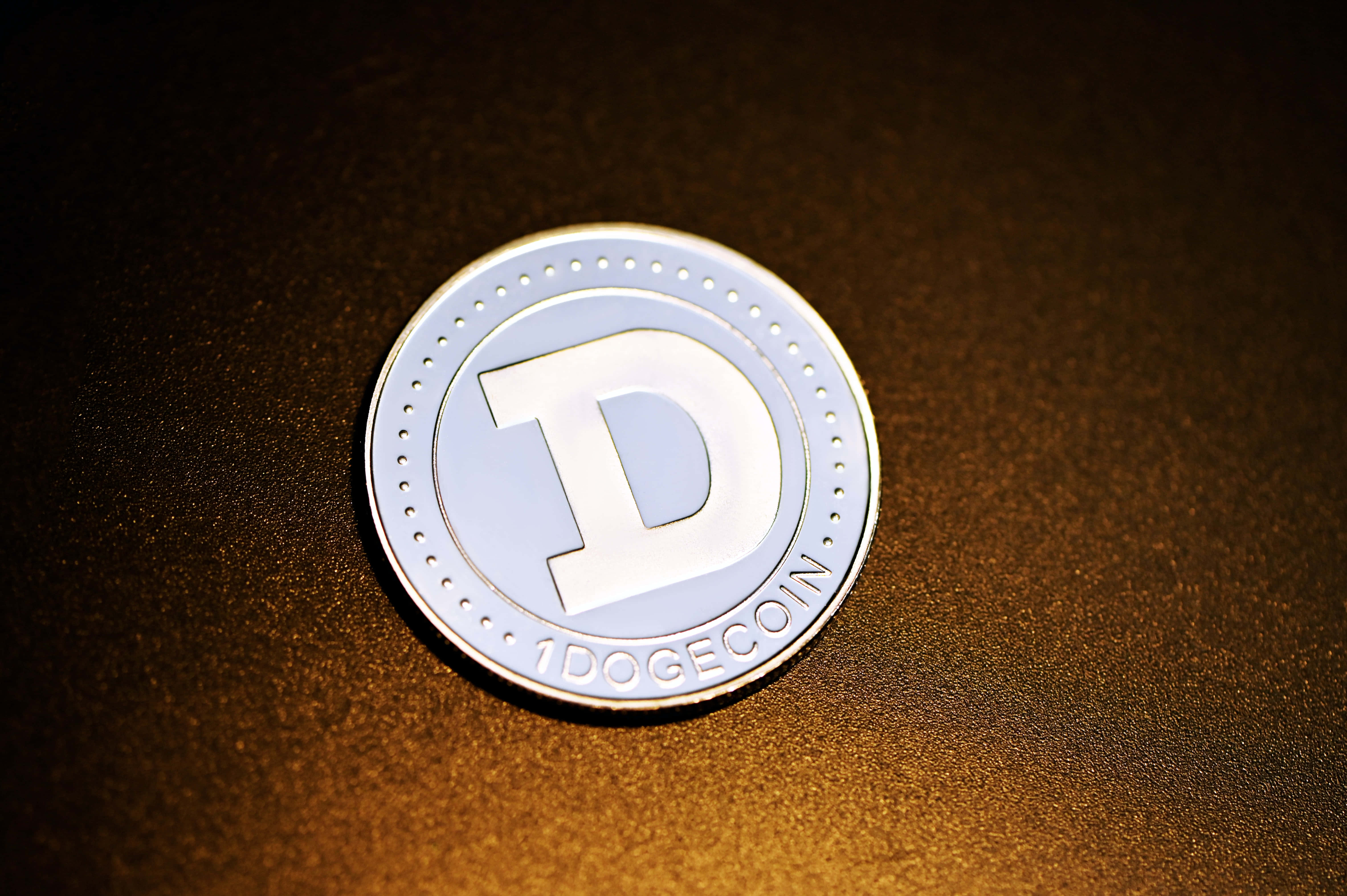 Dogecoin 桌布 壁紙 — 一枚閃亮的銀色Dogecoin代幣放在溫暖燈光下的紋理金色表面上。