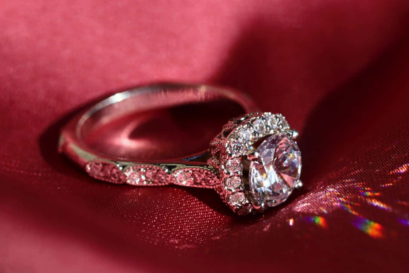 Ring 桌布 壁紙 — 一枚閃爍的鑽石訂婚戒指，採用光環鑲嵌，放置在濃郁的紅色天鵝絨布料上，帶有彩虹光折射。