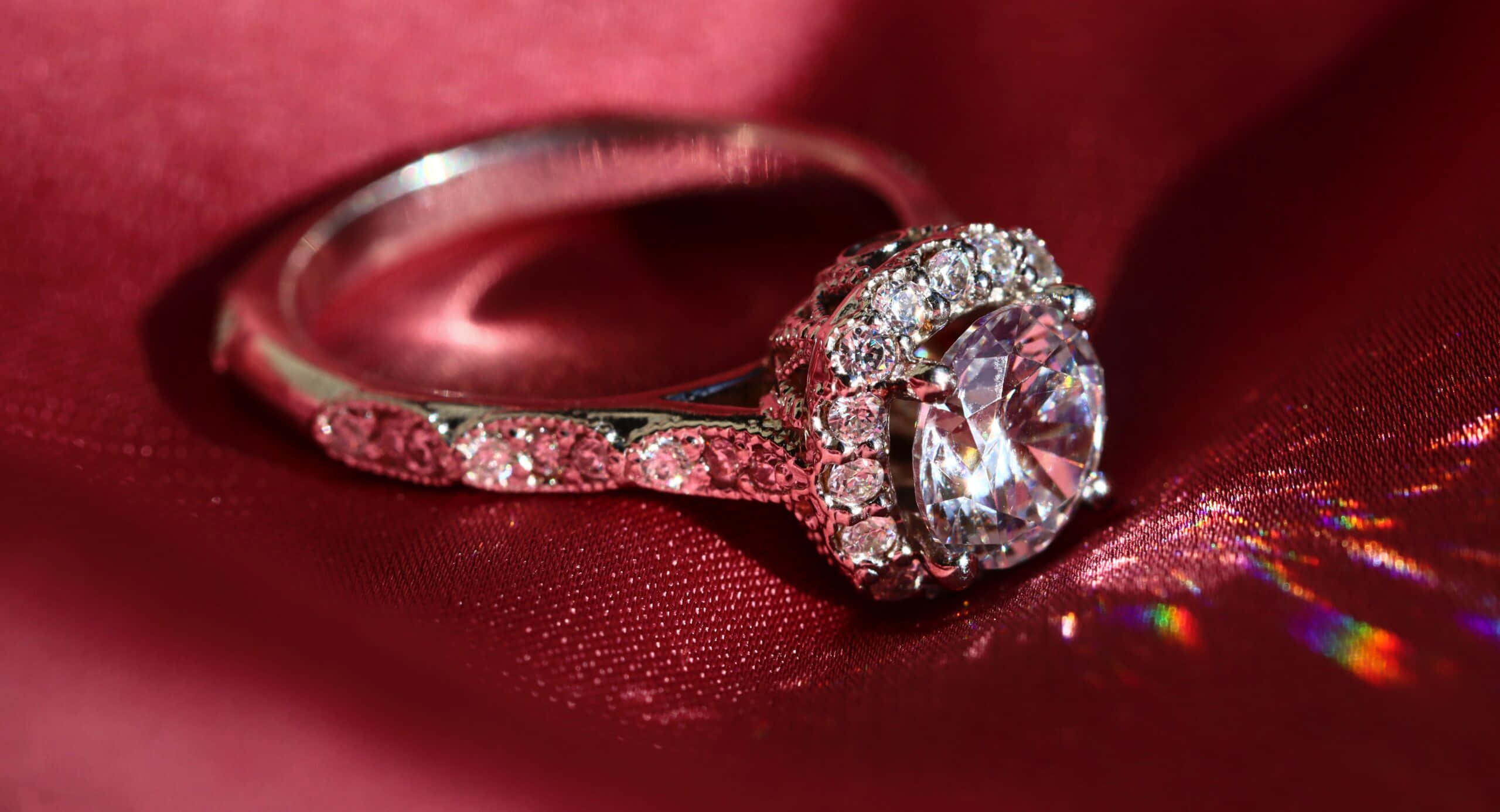 Wedding Rings 桌布 壁紙 — 一枚閃爍的鑽石訂婚戒指，採用密鑲鑲嵌，放置在豐富的紅色緞面布料上，帶有彩虹光反射。