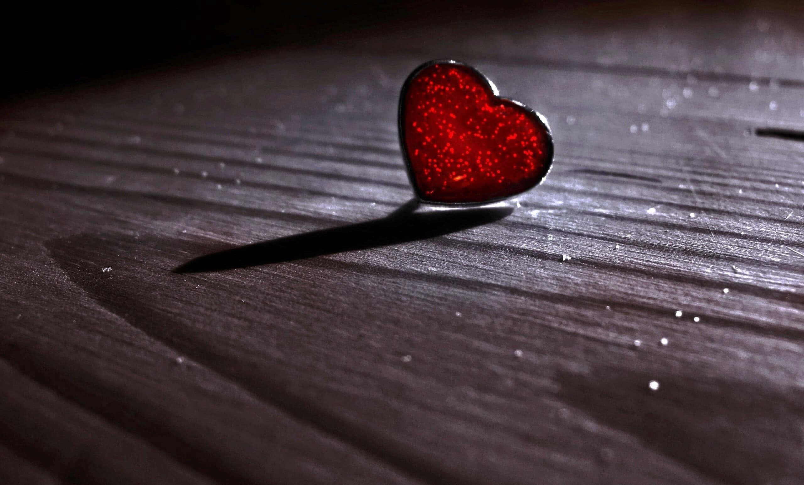 Red Heart 桌布 壁紙 — 一枚閃閃發光的紅色心形別針，在戲劇性的聚光燈下放在深色木質表面上。