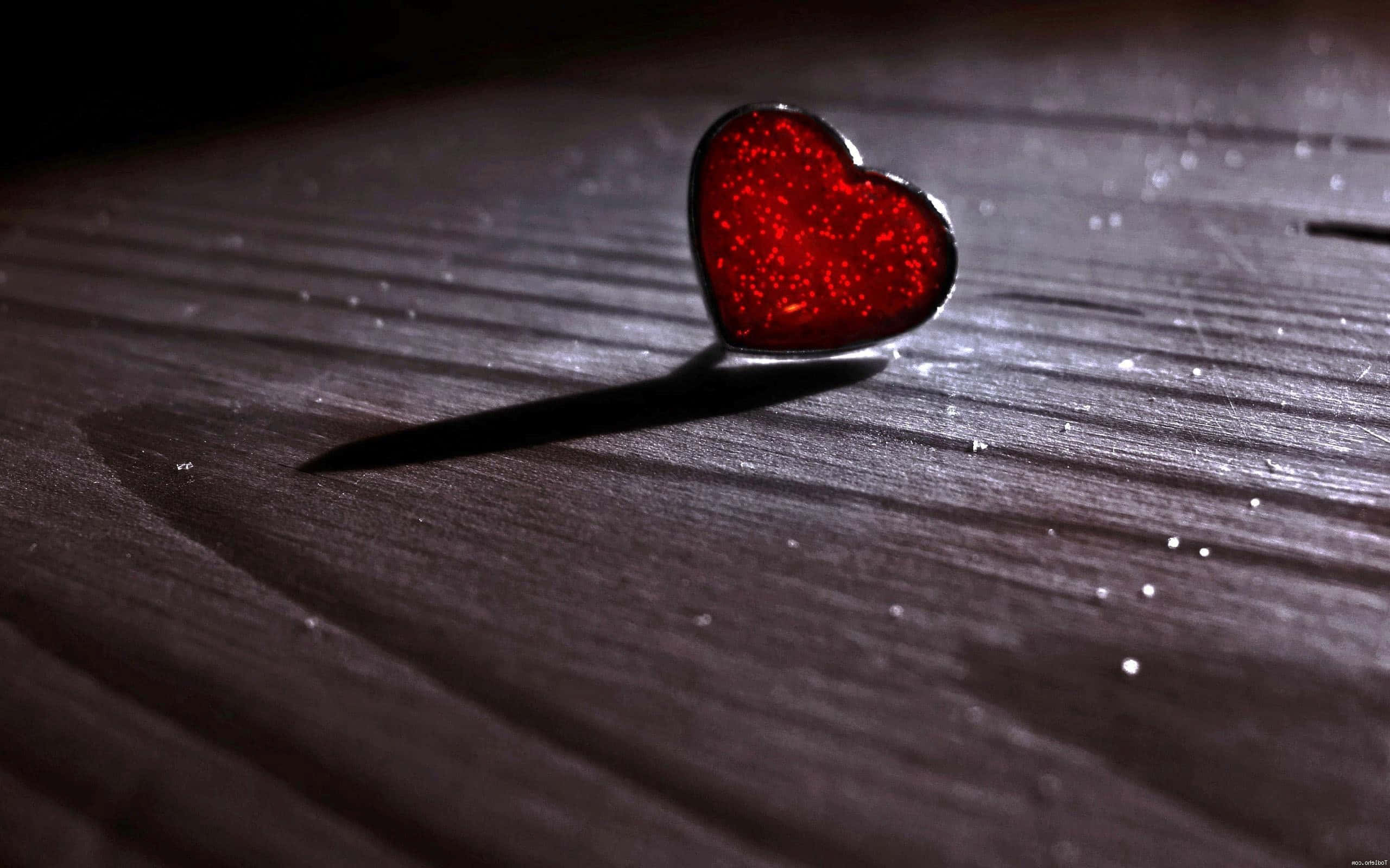 3d Heart 桌布 壁紙 — 一枚閃閃發光的紅色心形別針放在深色木質表面上，在戲劇性的燈光下投射出長長的陰影。