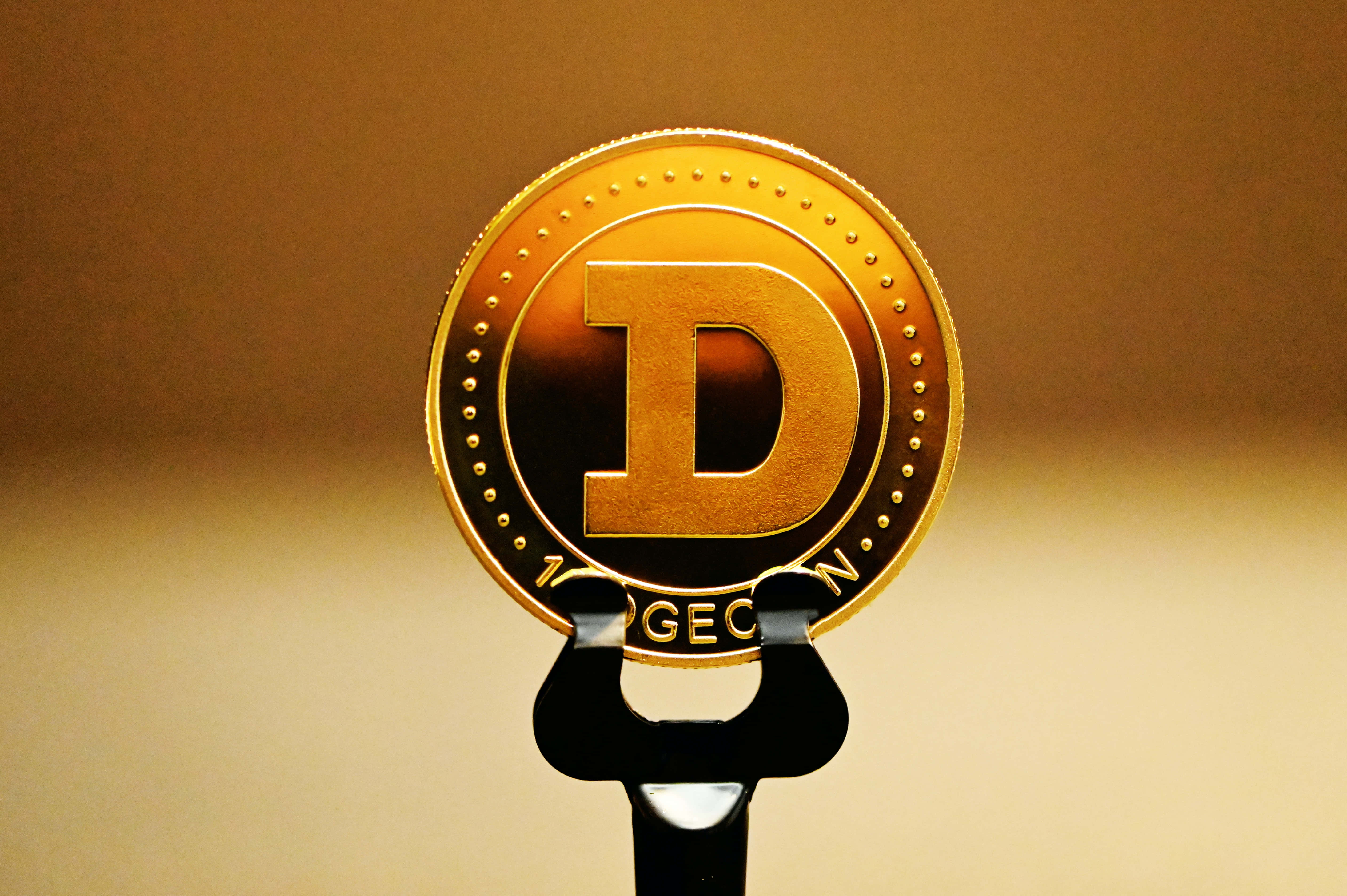 Dogecoin 桌布 壁紙 — 一枚閃閃發光的金幣，上面有一個大D標誌，放在黑色支架上，背景溫暖模糊。