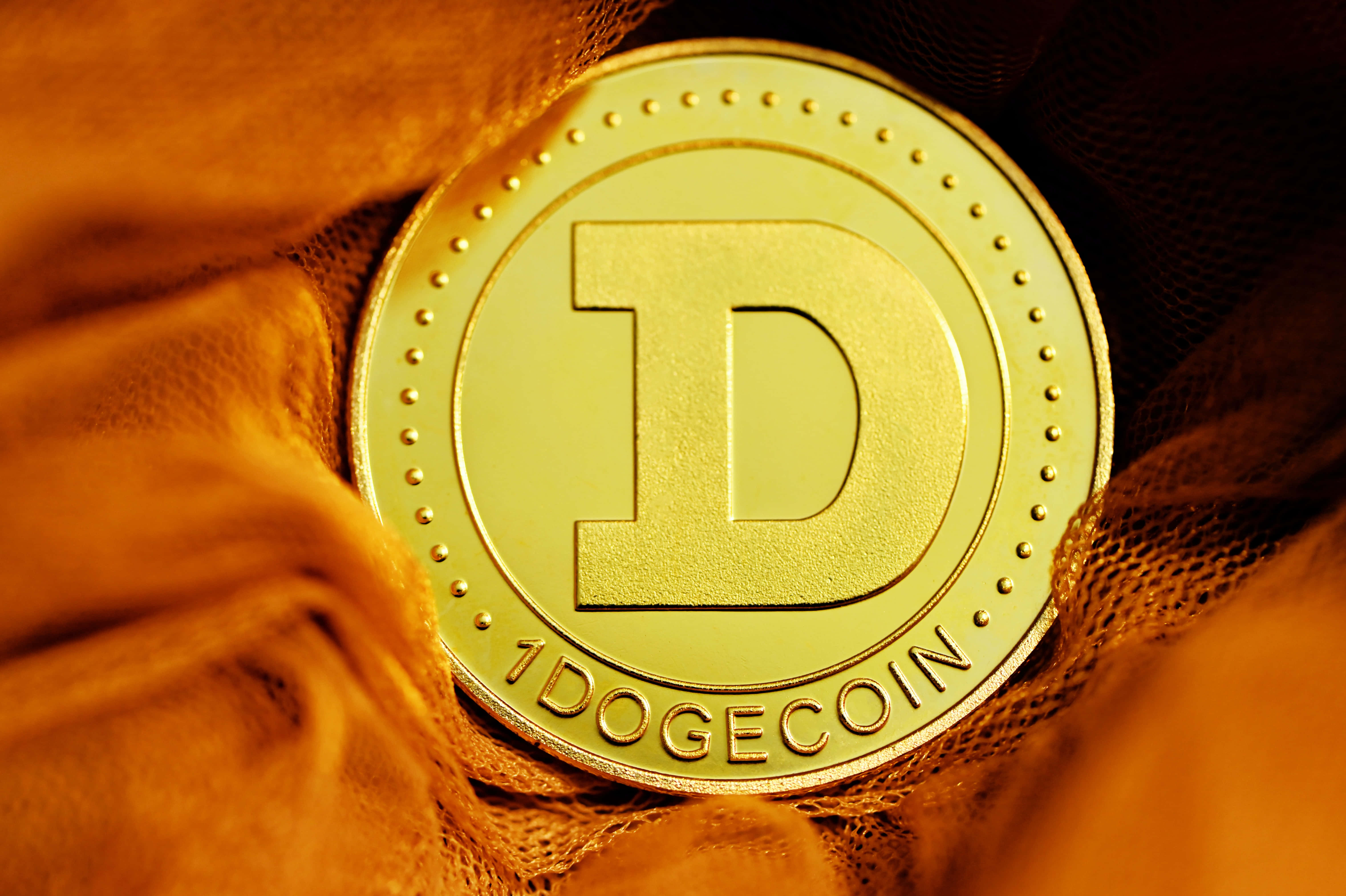 Dogecoin 桌布 壁紙 — 一枚閃閃發光的金色狗狗幣放在溫暖燈光下的紋理橙色布料上。