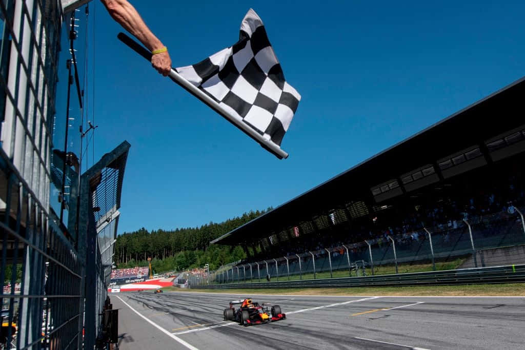 Chequered Flag 桌布 壁紙 — 一架一級方程式賽車越過終點線，同時有一隻手在晴朗的藍天下揮動格仔旗。