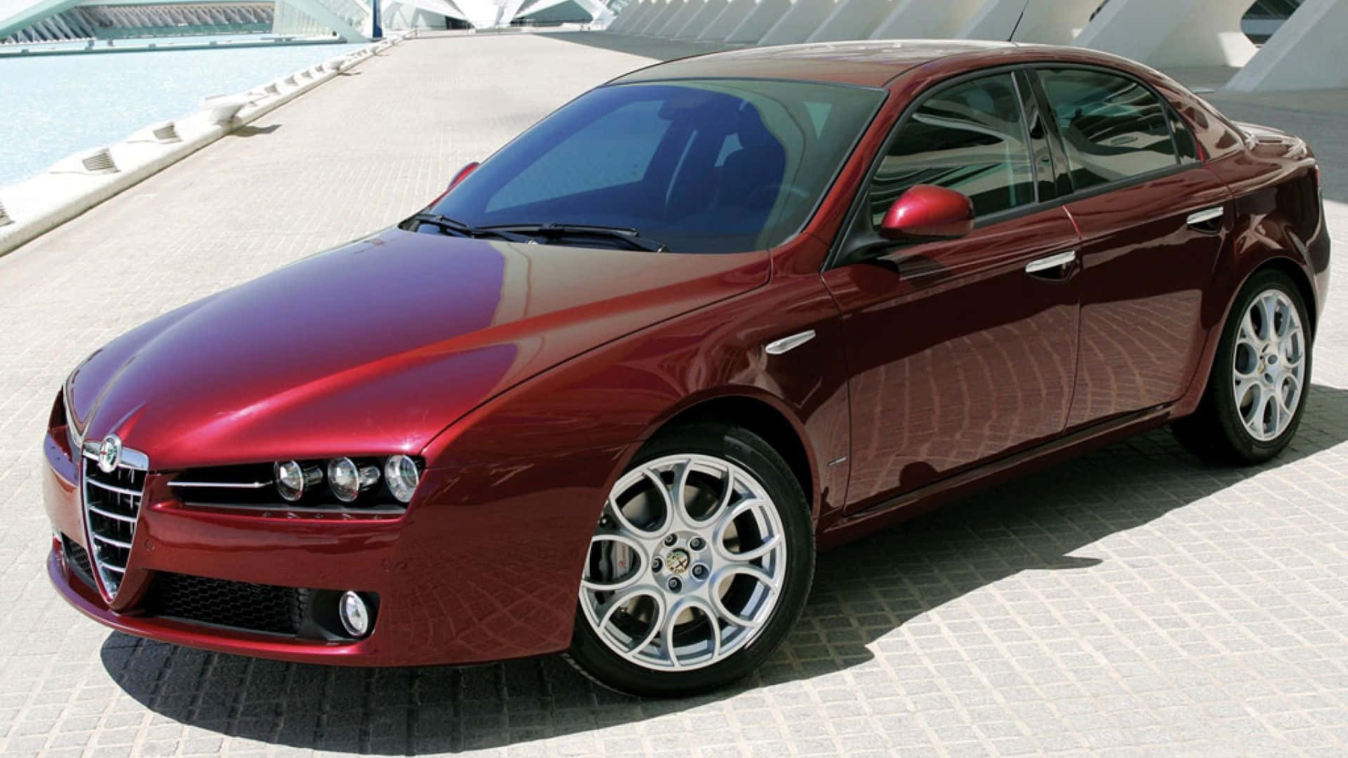 Alfa Romeo 159 桌布 壁紙 — 一架亮紅色Alfa Romeo房車停泊在瓷磚廣場上，旁邊是現代建築和水景。
