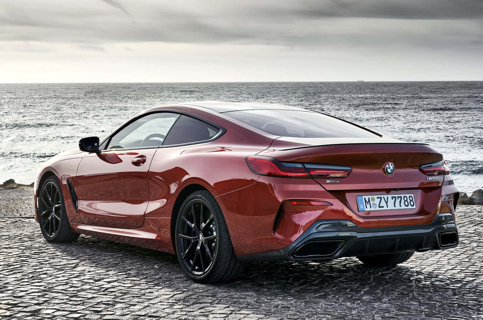 Bmw 8 Series 桌布 壁紙 — 一架亮紅色BMW 8系轎跑車停泊在鵝卵石海岸上，背後是廣闊的海洋和陰天。