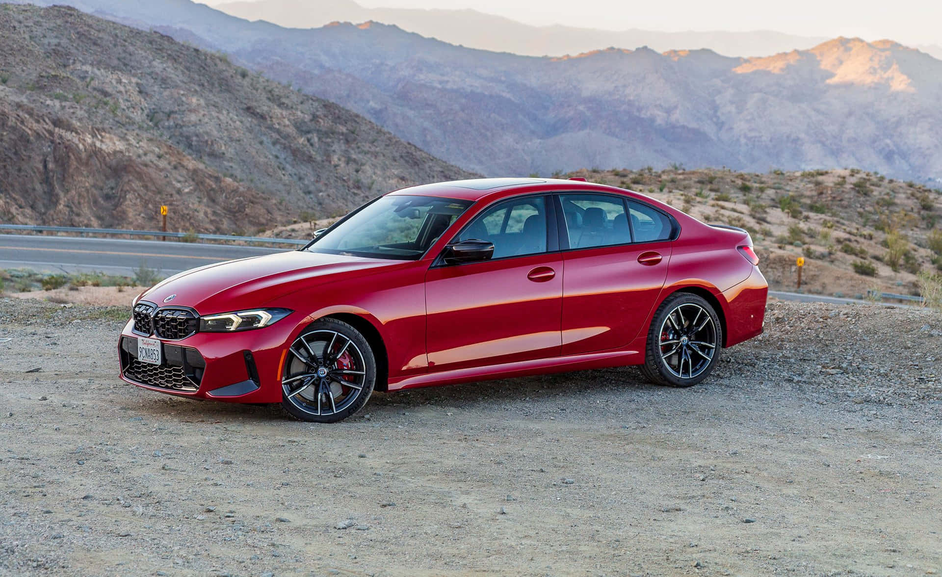 Bmw 3 Series 桌布 壁紙 — 一架亮紅色BMW M3房車停在沙漠公路旁的碎石上，崎嶇的山脈沐浴在金黃色的日落光芒中。