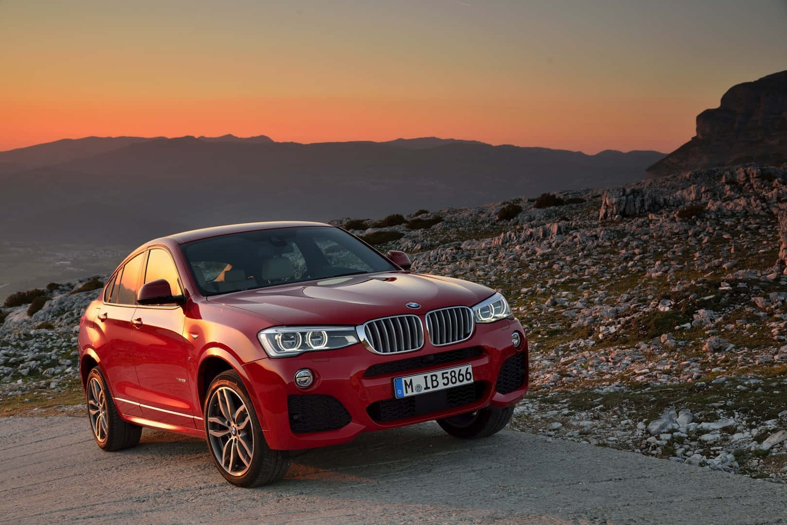 Bmw X4 桌布 壁紙 — 一架亮紅色BMW X4 SUV停泊在日落時分的山路，天空橙黃，地形岩石。