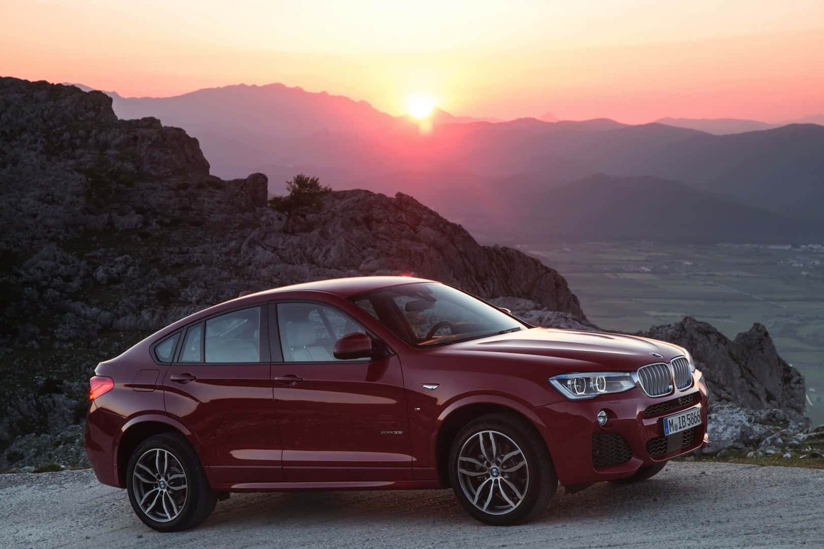 Bmw X4 桌布 壁紙 — 一架亮紅色BMW X6停泊在岩石觀景點，夕陽在遠山後落下，天空染上溫暖的橙色和粉紅色調。