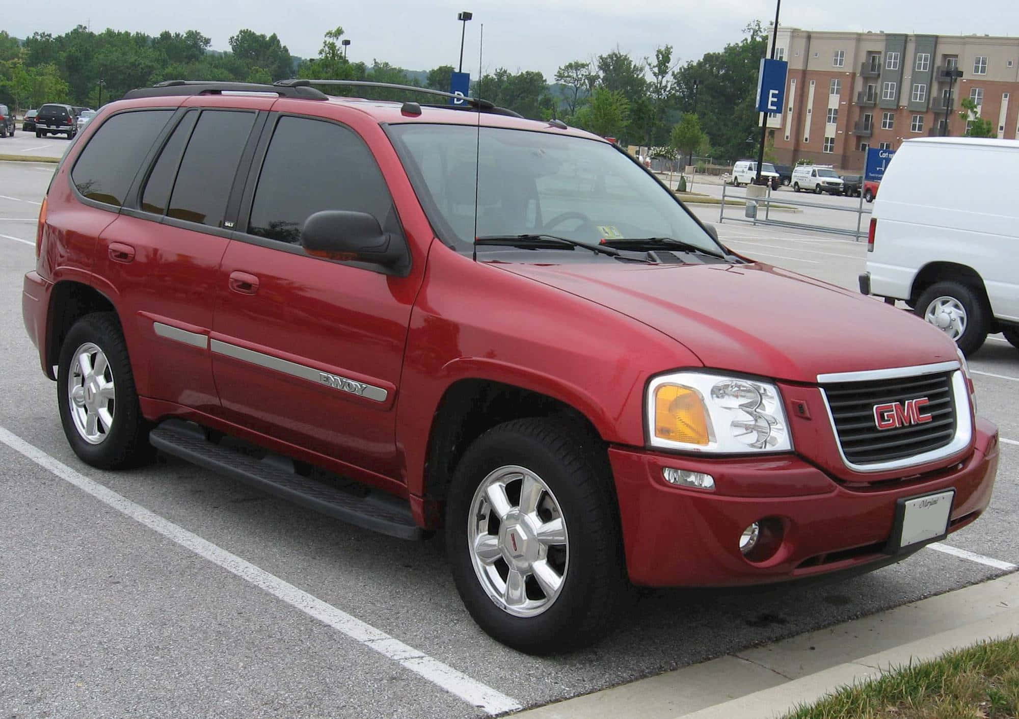Gmc Envoy 桌布 壁紙 — 一架亮紅色GMC Envoy SUV停泊在鋪砌的停車場，背景有樹木和公寓大樓。