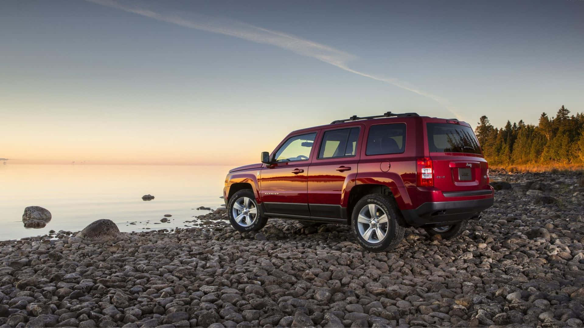 Jeep Patriot 桌布 壁紙 — 一架亮紅色SUV停泊在日落時分的岩石湖岸邊，平靜的水面和遠處的樹木在柔和漸變的天空下。