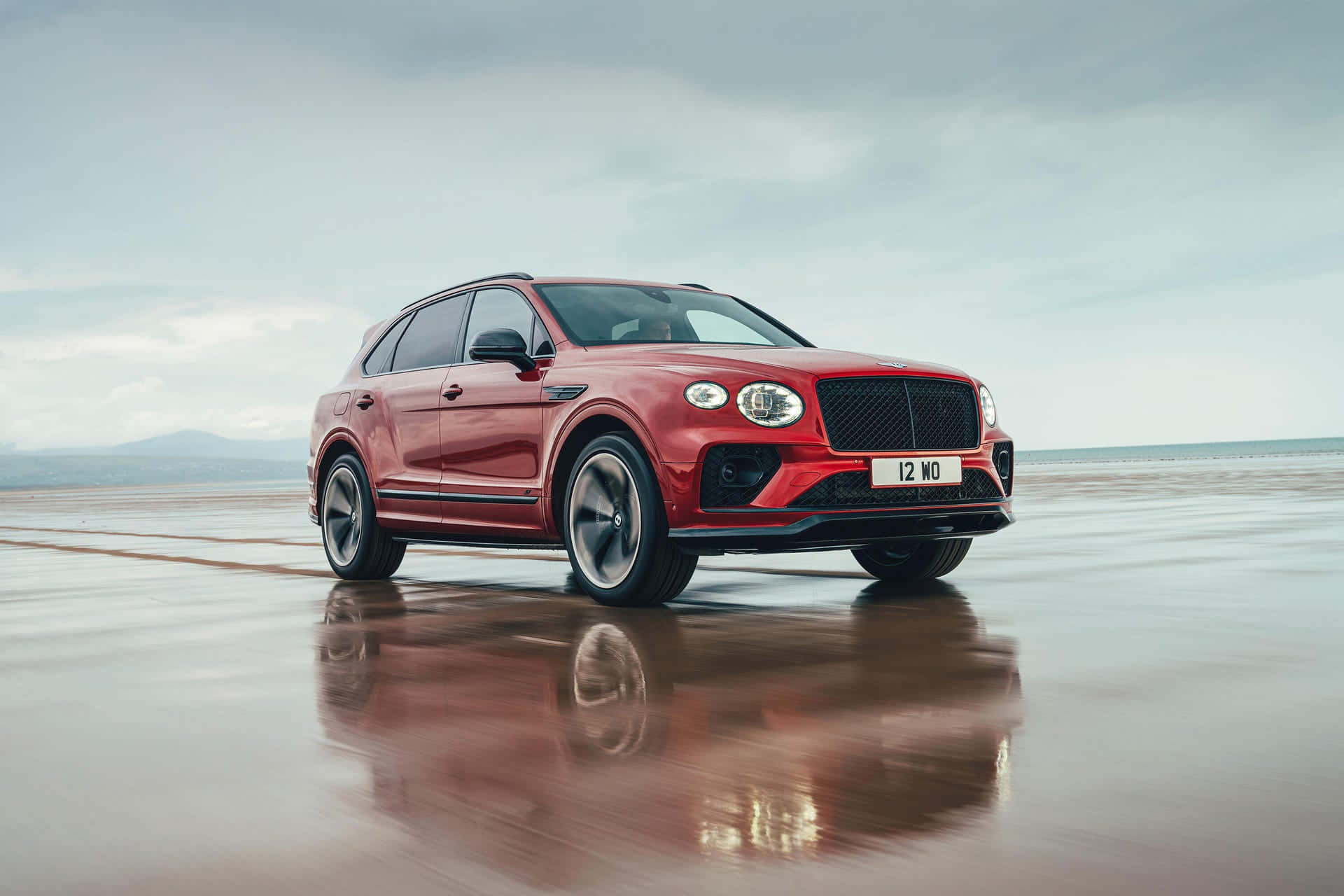 Bentley Bentayga 桌布 壁紙 — 一架亮紅色嘅 Bentley Bentayga SUV 停喺濕嘅沙灘上，喺多雲嘅天空下反射出佢流線型嘅外形。