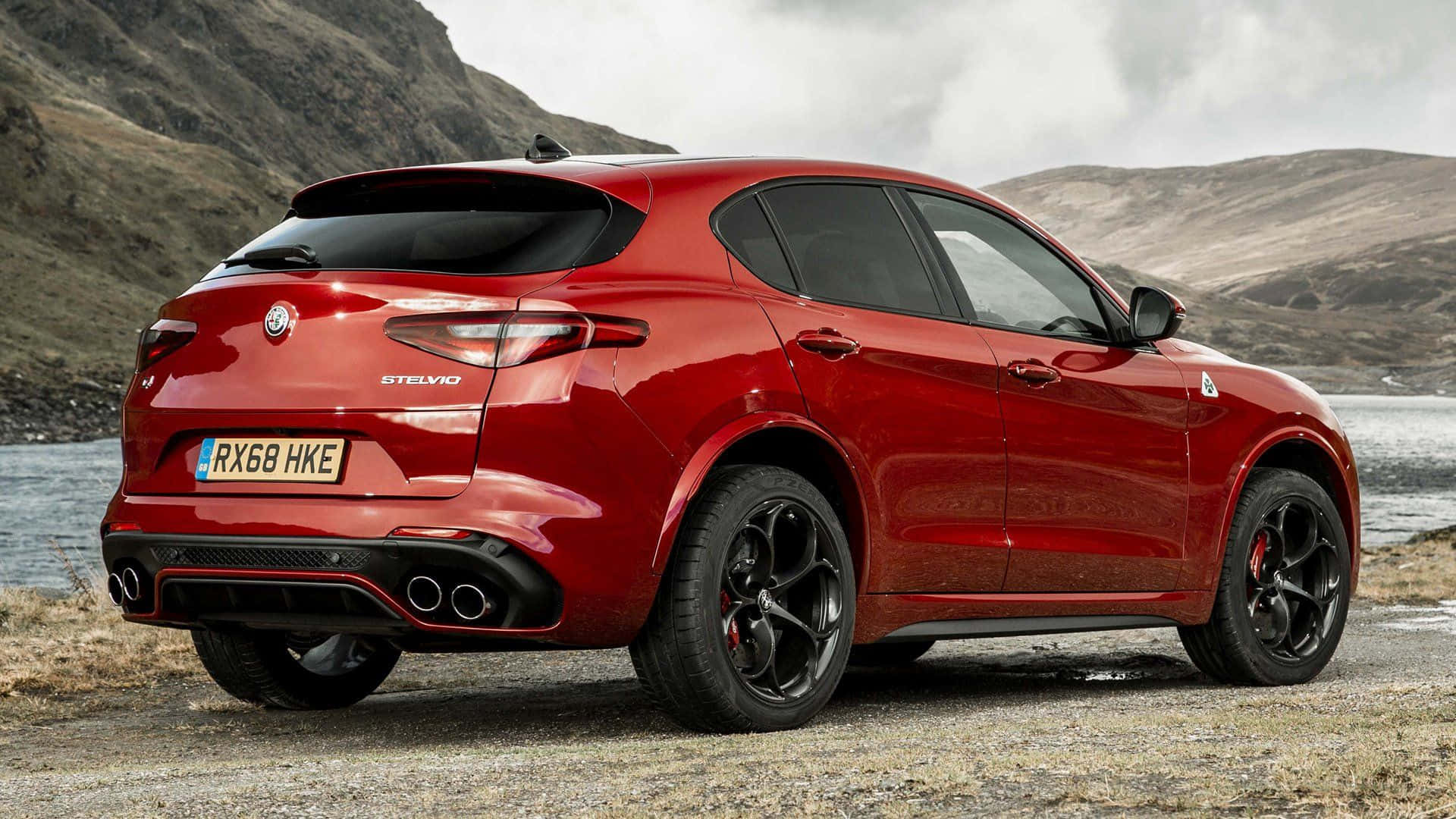 Alfa Romeo Stelvio 桌布 壁紙 — 一架亮紅色嘅Alfa Romeo Stelvio SUV停泊喺碎石岸邊，旁邊係平靜嘅湖泊，背景有崎嶇嘅山丘，天空多雲。
