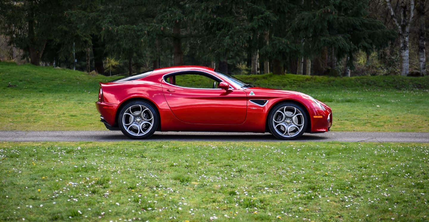 Alfa Romeo 8c Competizione 桌布 壁紙 — 一架亮紅色嘅Alfa Romeo跑車泊喺碎石路旁邊，隔籬係有白色花朵點綴嘅草地。