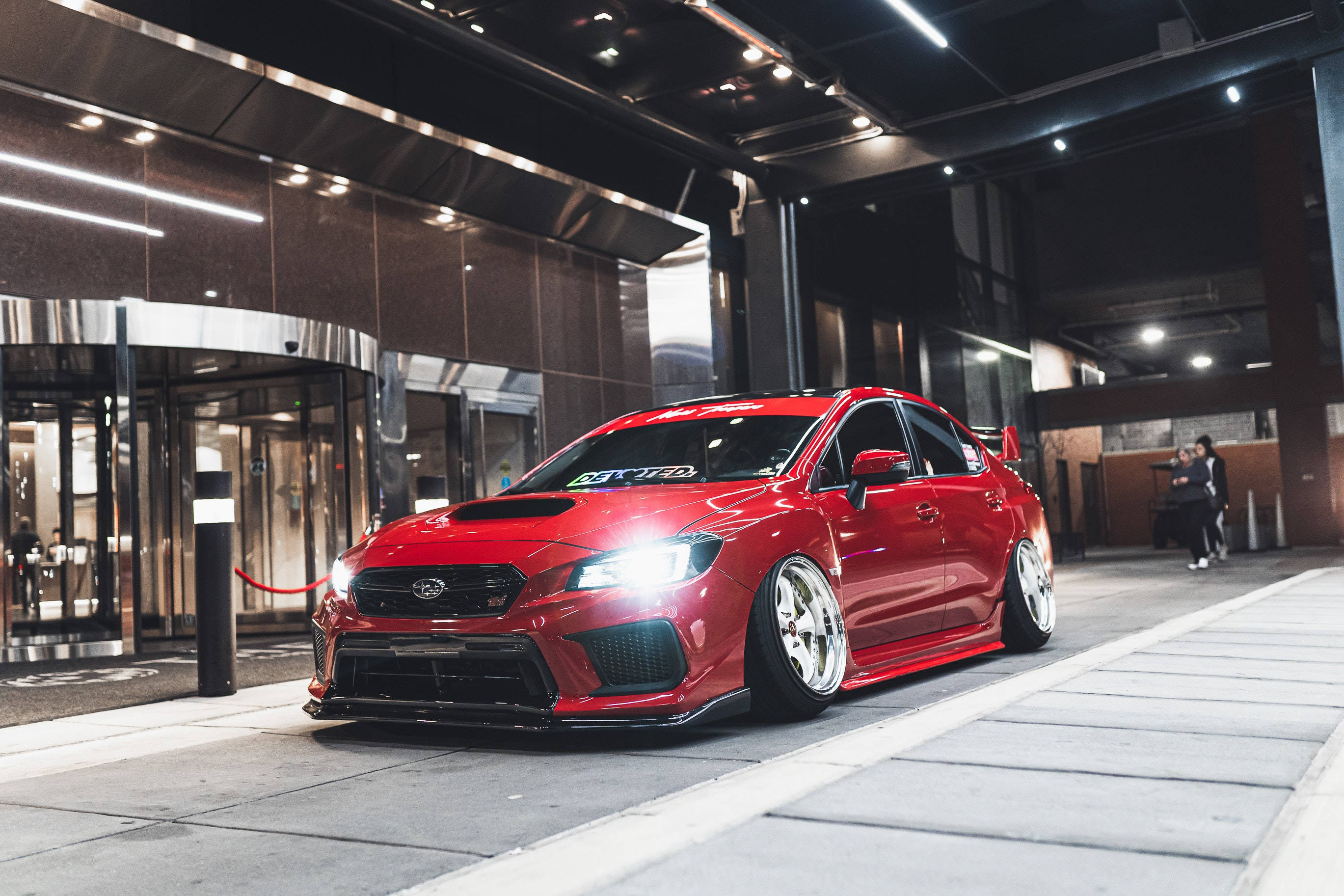 subaru 桌布 壁紙 — 一架亮紅色改裝Subaru WRX STI夜晚停喺現代建築物外面，車頭燈著住。
