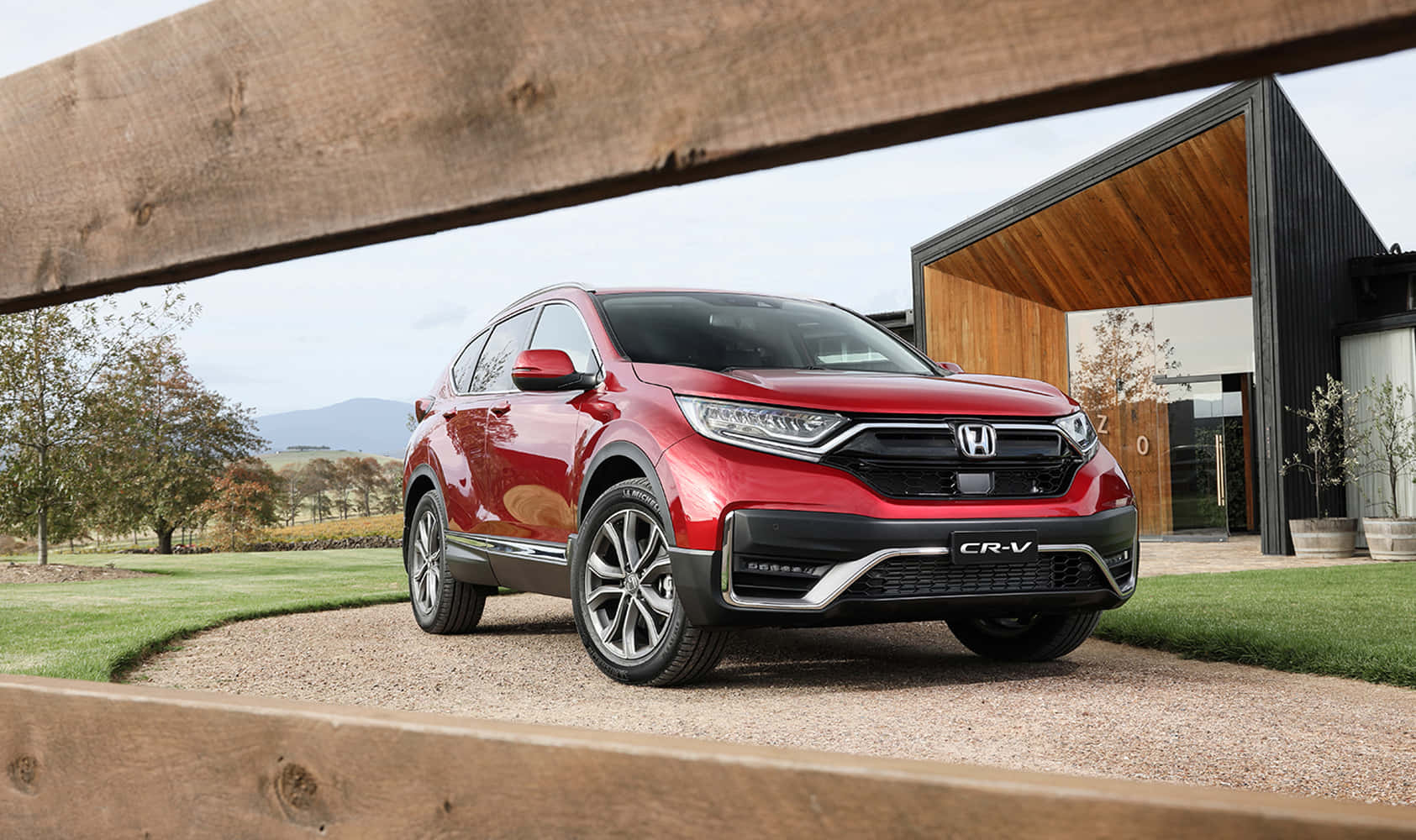 Honda Cr-v 桌布 壁紙 — 一架亮紅色本田CR-V SUV停泊在碎石車道上，車道由木樑框起，背景是一座現代房屋和連綿起伏的山丘。