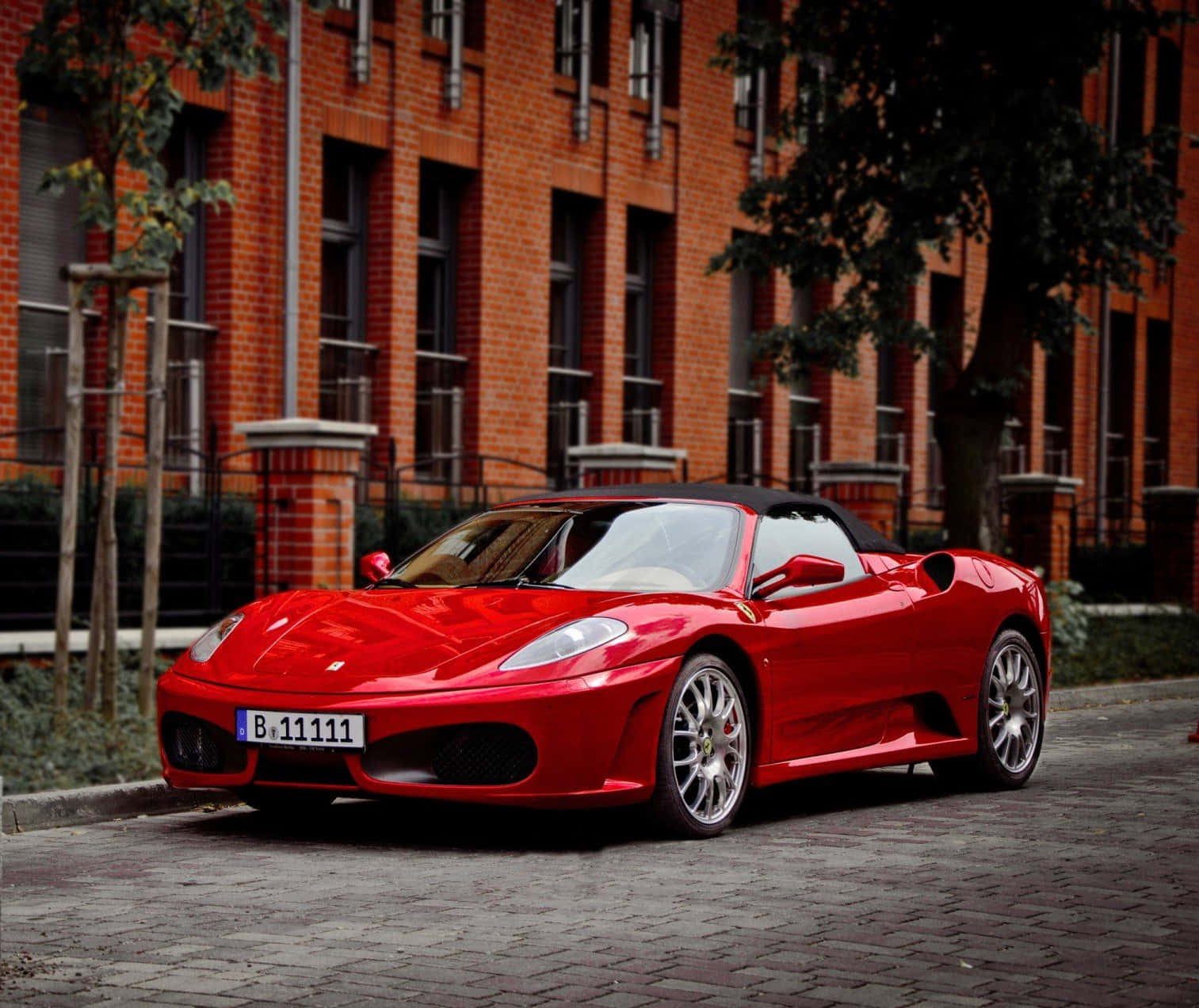 Ferrari F430 桌布 壁紙 — 一架亮紅色法拉利 Spider 停泊在鵝卵石街道上，前面係一棟有樹木嘅磚建築。