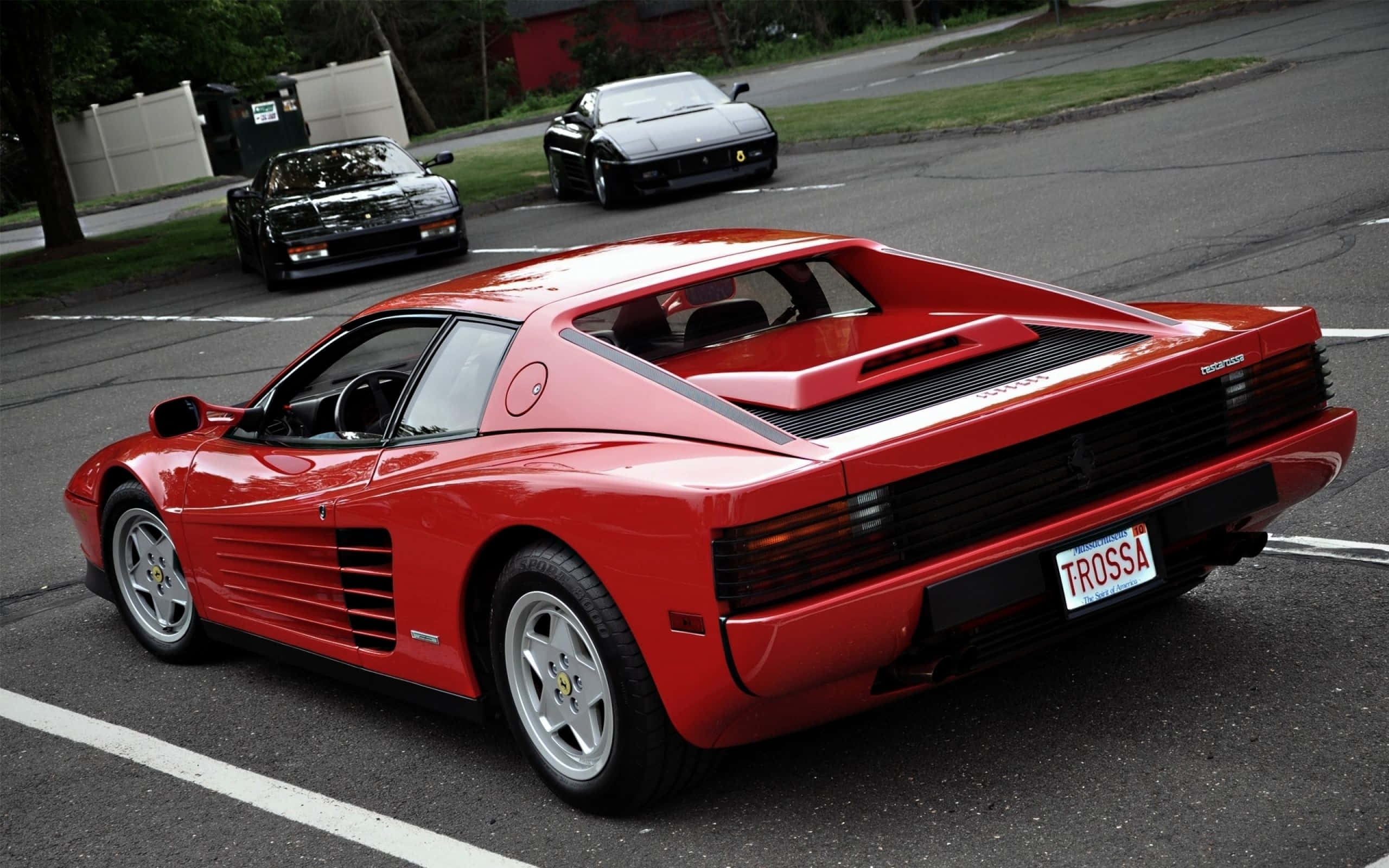 Ferrari Testarossa 桌布 壁紙 — 一架亮紅色法拉利 Testarossa 停泊在停車場，背景有兩輛黑色跑車。