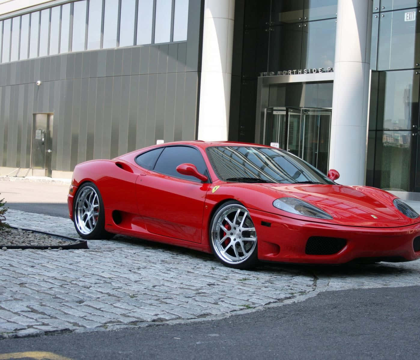 Ferrari 360 Modena 桌布 壁紙 — 一架亮紅色法拉利360跑車停泊在現代玻璃建築前的鵝卵石車道上。