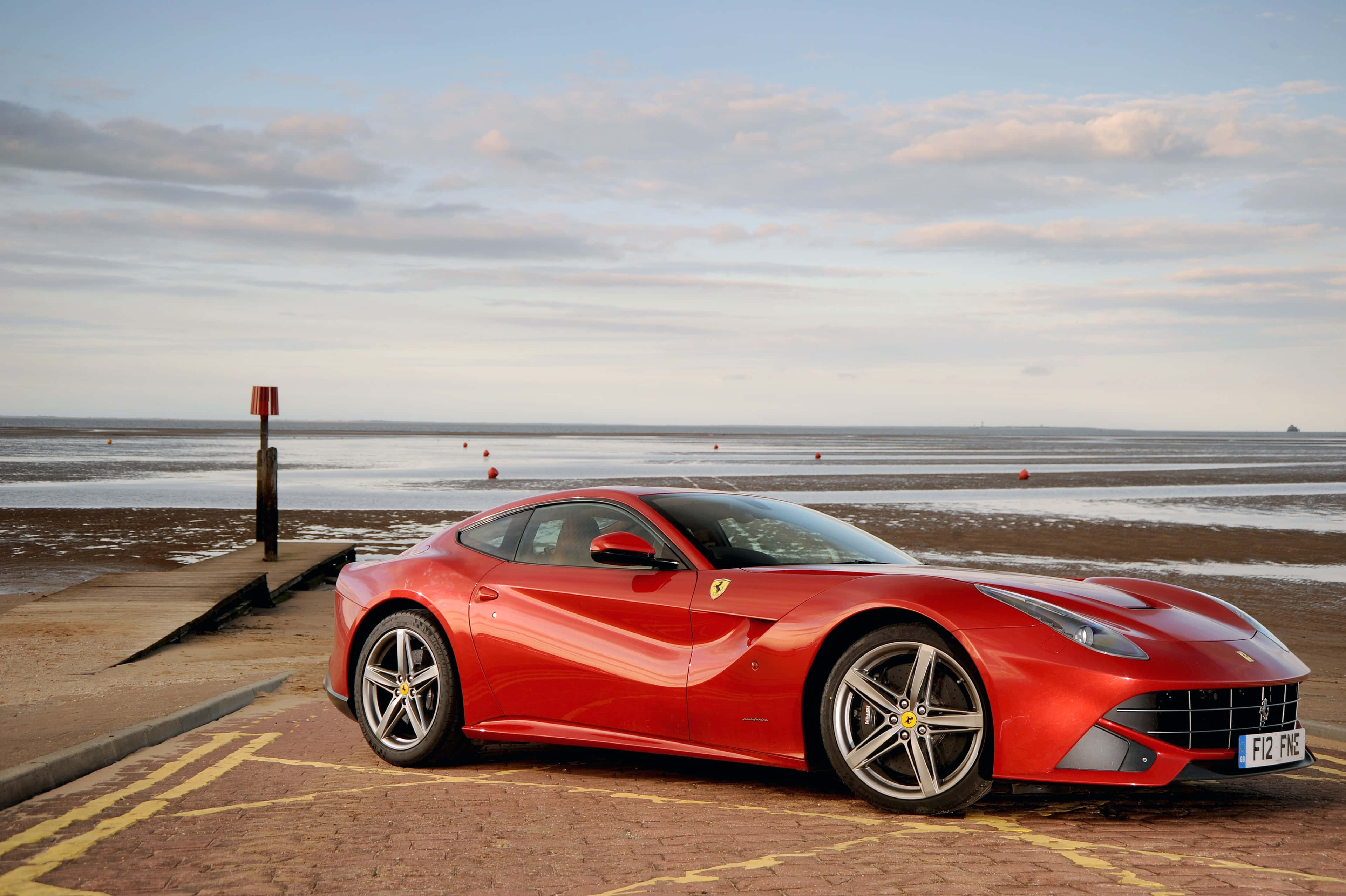 Ferrari F12 Berlinetta 桌布 壁紙 — 一架亮紅色法拉利F12停泊在磚砌碼頭上，背景是日落時分的平靜海面和散落的浮標。