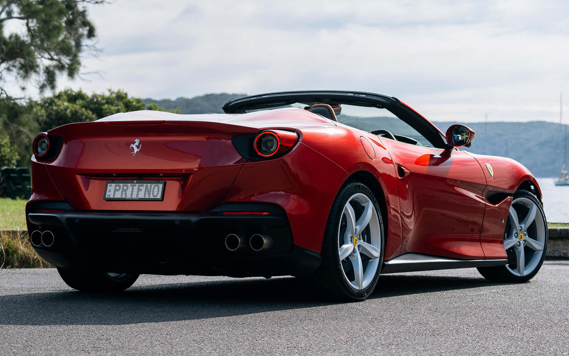 Ferrari Portofino 桌布 壁紙 — 一架亮紅色法拉利Portofino開篷跑車停泊在沿海道路上，背景有樹木和水面。
