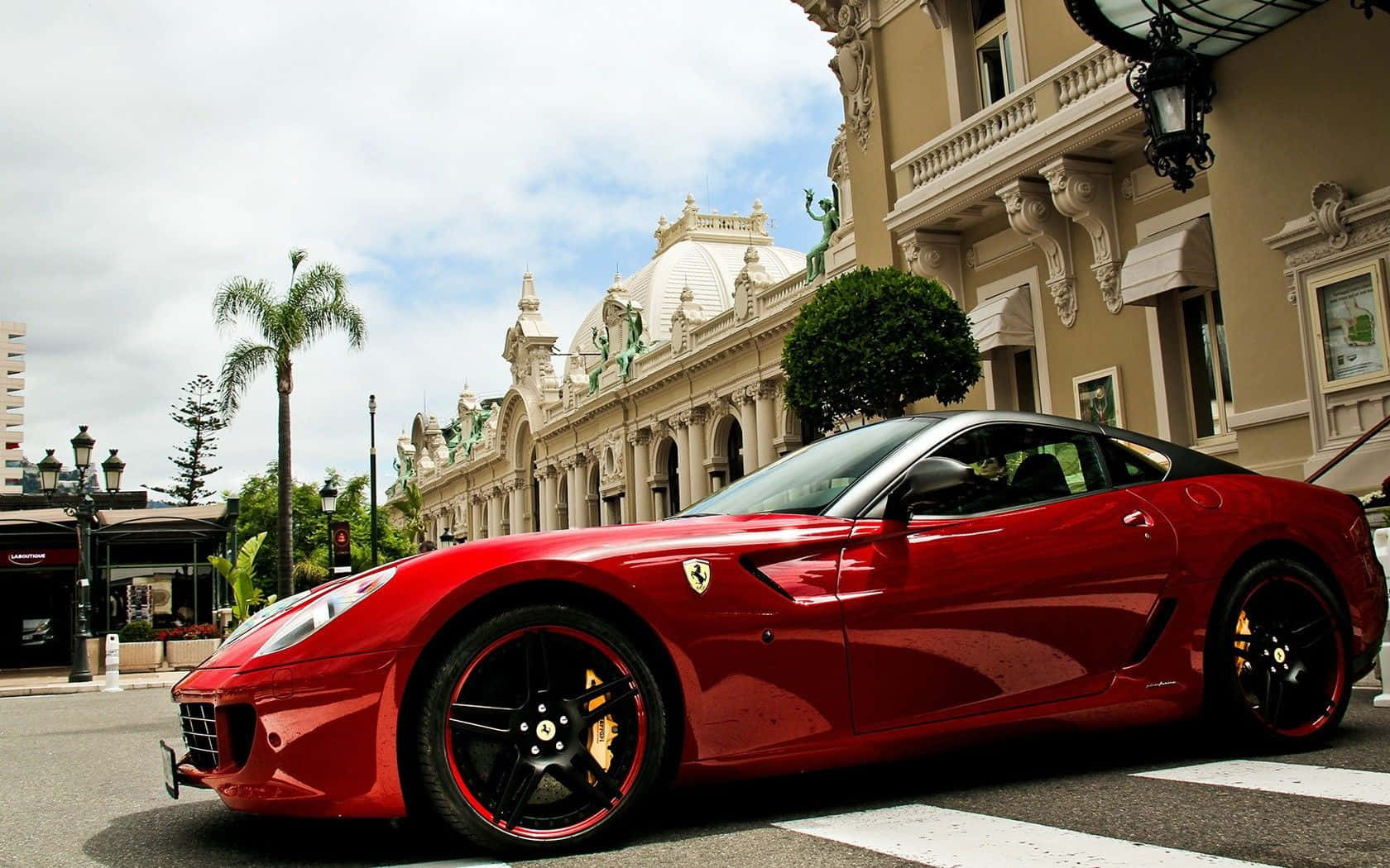 Ferrari 599 Gtb Fiorano 桌布 壁紙 — 一架亮紅色法拉利跑車停泊在街上，旁邊係一棟有棕櫚樹嘅裝飾性米色建築，天空局部多雲。