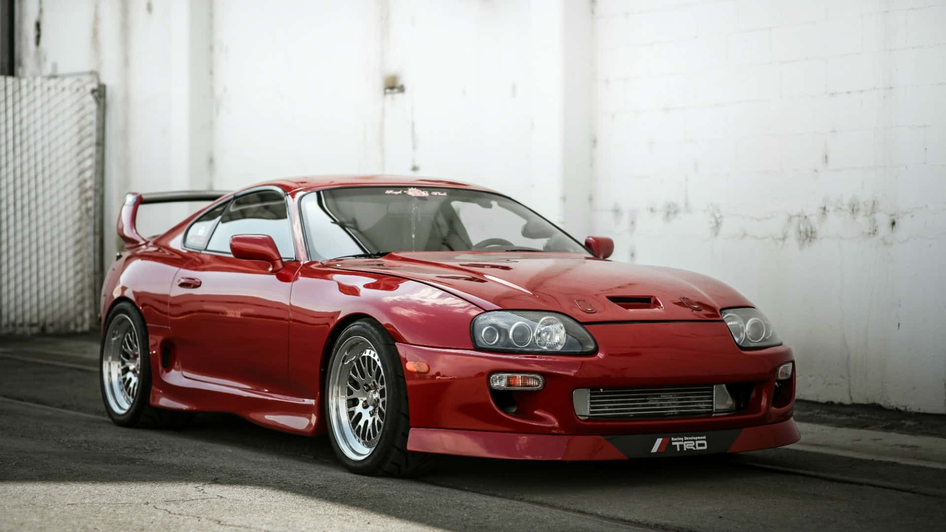 Supra 桌布 壁紙 — 一架亮紅色豐田Supra跑車，帶有後擾流板，停泊在風化的白色混凝土牆旁。