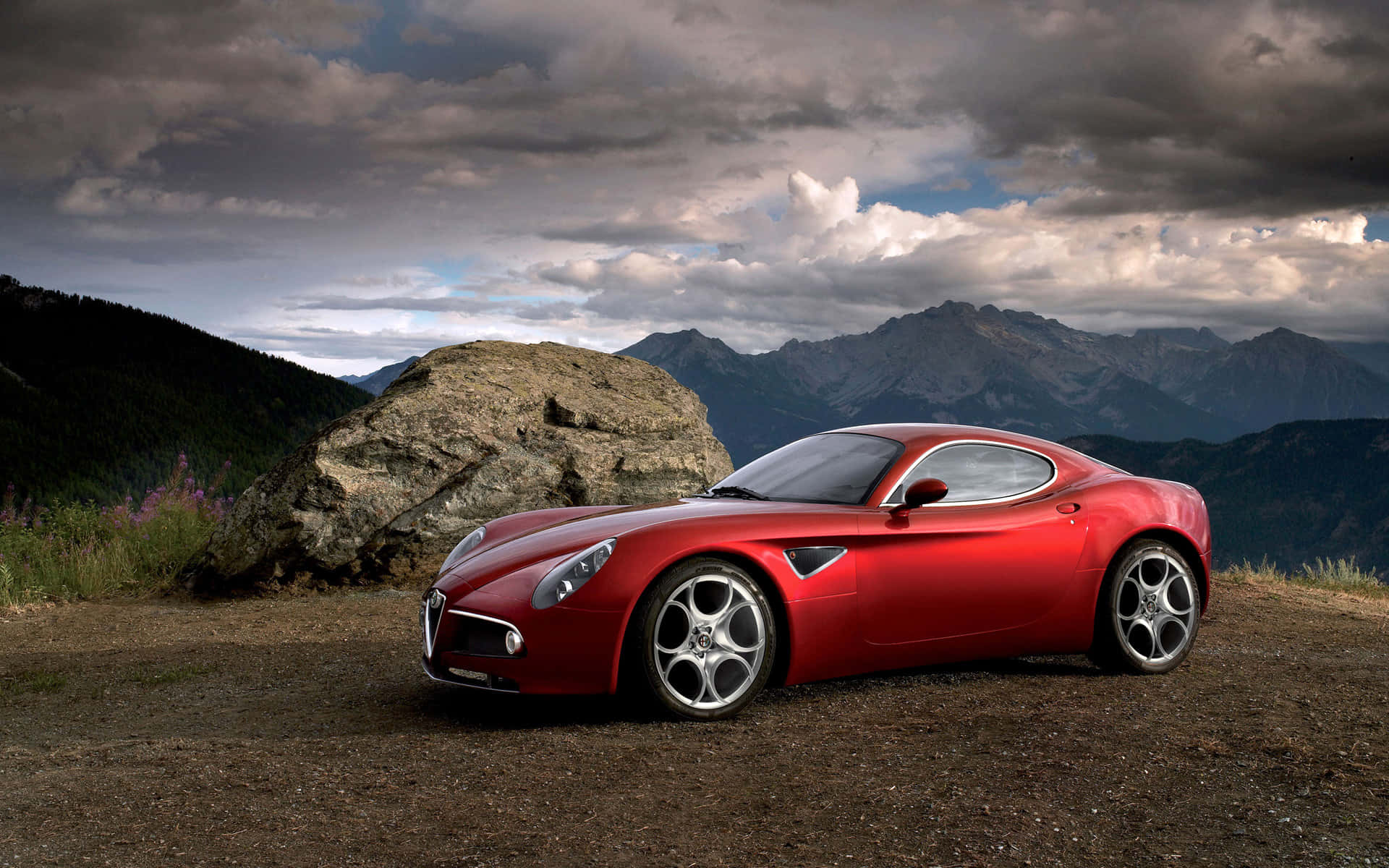 Alfa Romeo 8c Competizione 桌布 壁紙 — 一架亮紅色跑車停泊在山脊上，背景是戲劇性的多雲天空和崎嶇的山峰。