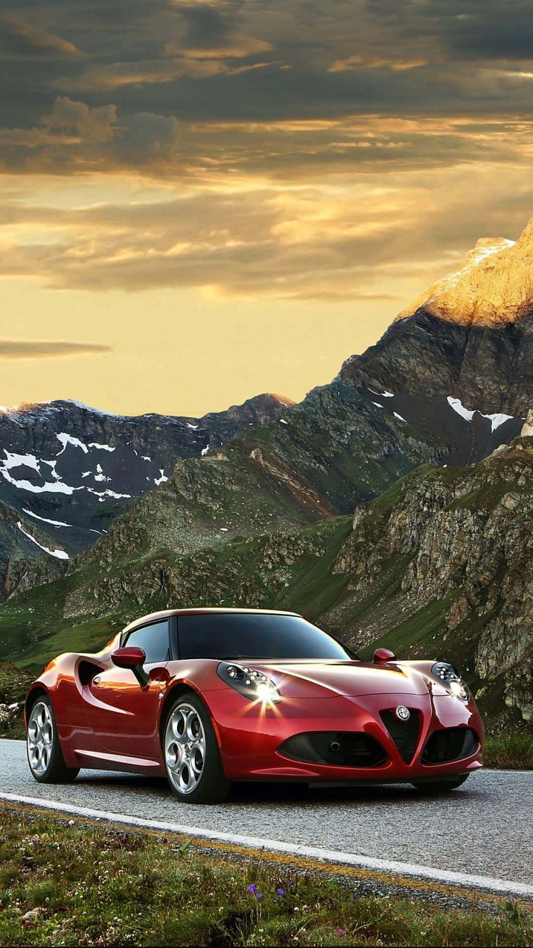 Alfa Romeo 4c 桌布 壁紙 — 一架亮紅色跑車在日落時分行駛在山路上，背景是白雪覆蓋的山峰和金色的雲彩。
