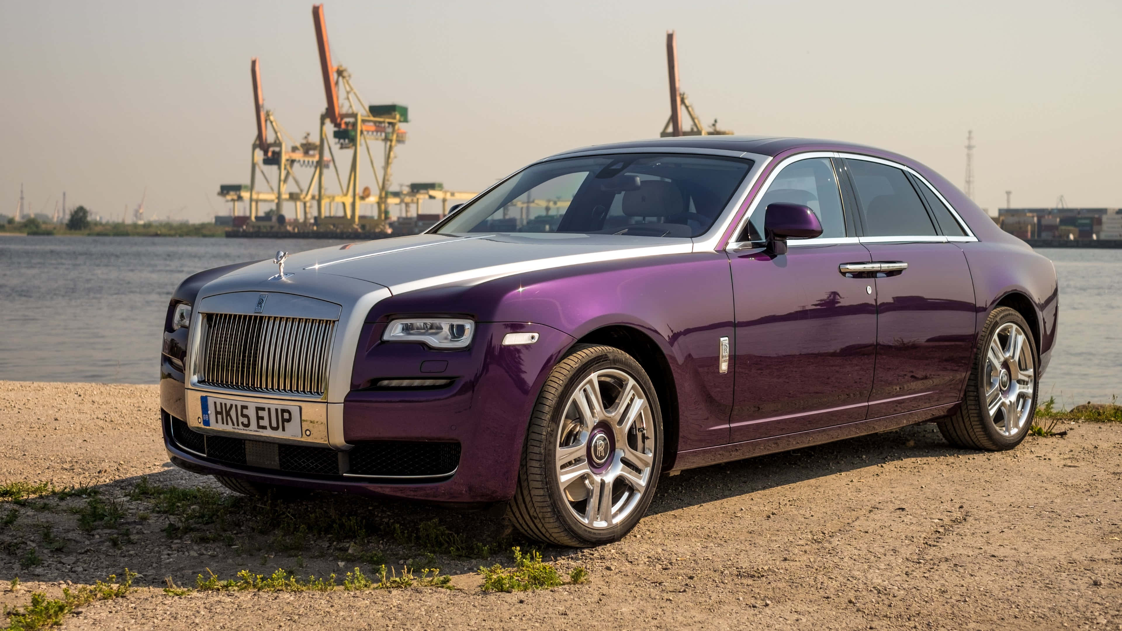 Rolls Royce 桌布 壁紙 — 一架亮紫色嘅勞斯萊斯 Ghost 停泊喺碎石灘旁邊，水面有工業起重機喺朦朧嘅背景中。