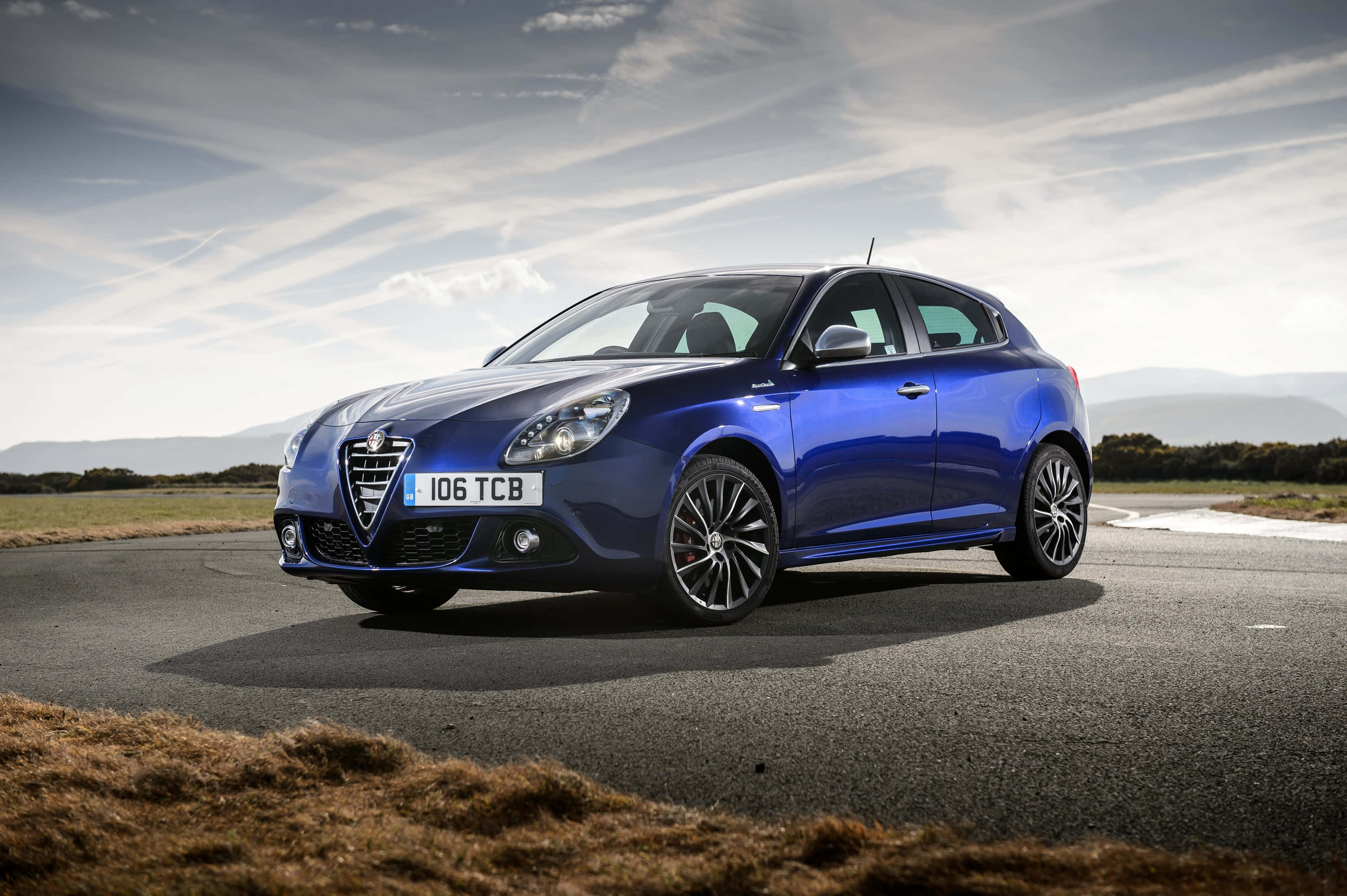 Alfa Romeo Giulietta 桌布 壁紙 — 一架亮藍色愛快羅密歐Giulietta停泊在蜿蜒道路上，遠方山丘在陰天下。