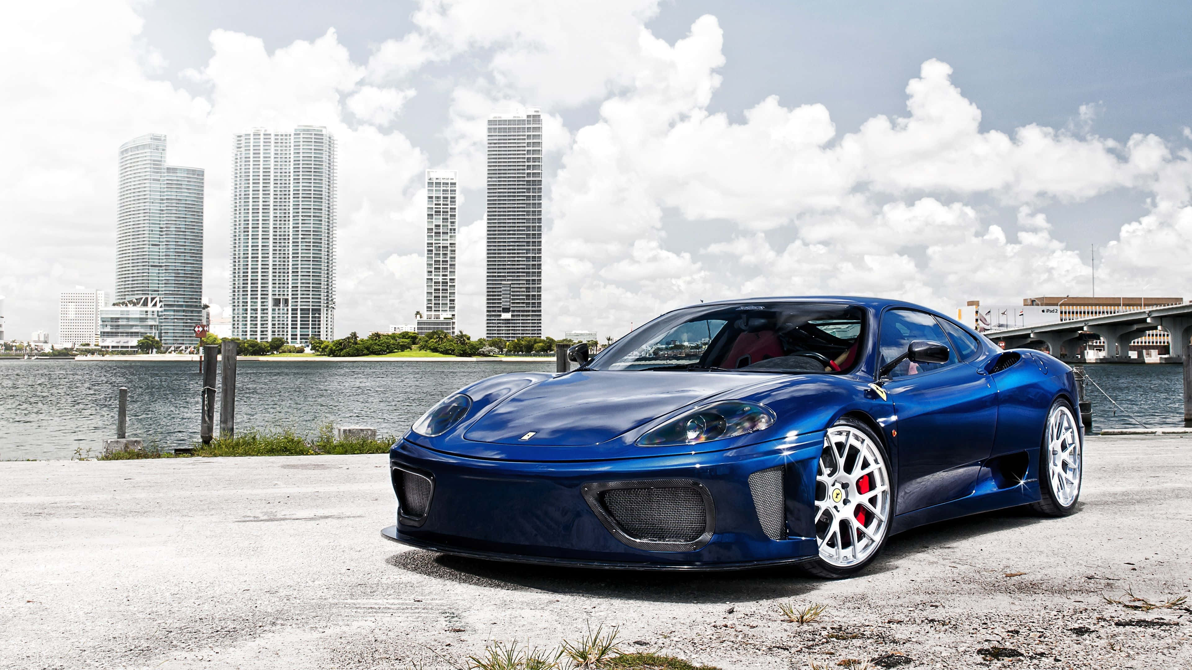 Ferrari 360 Modena 桌布 壁紙 — 一架亮藍色法拉利跑車停泊在海濱碼頭，背景是現代摩天大樓和橋樑，天空部分多雲。