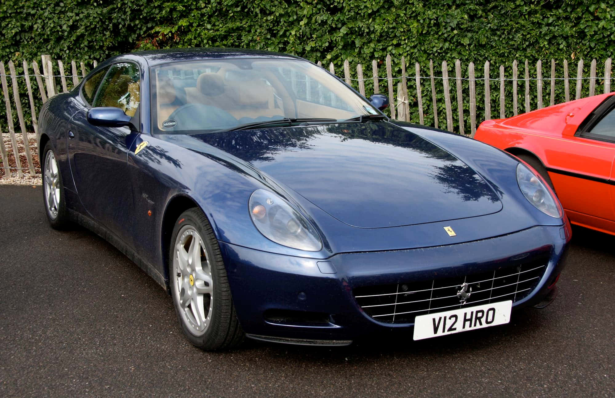 Ferrari 612 Scaglietti 桌布 壁紙 — 一架亮藍色法拉利跑車停泊在瀝青路上，旁邊有一輛紅色汽車，後面有綠色樹籬。