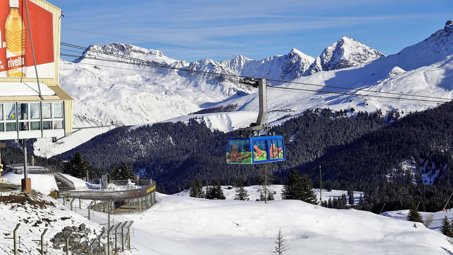 Ski Resort 桌布 壁紙 — 一架亮藍色的纜車在晴朗的藍天下載著乘客越過積雪的山脈，並可看到滑雪度假村建築。