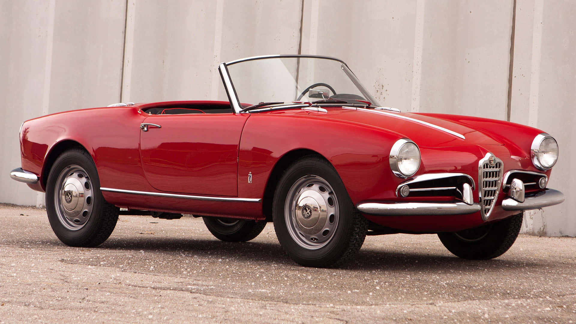 Alfa Romeo Spider 桌布 壁紙 — 一架亮面紅色經典愛快羅密歐Spider開篷車停泊在碎石上，背景是波浪形金屬牆。