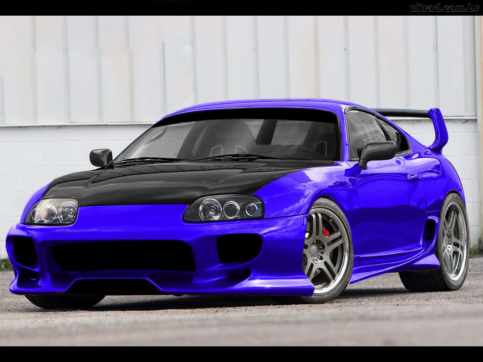 Supra 桌布 壁紙 — 一架亮面紫色和黑色豐田Supra跑車，帶有後尾翼，停在白色牆前的瀝青路面上。
