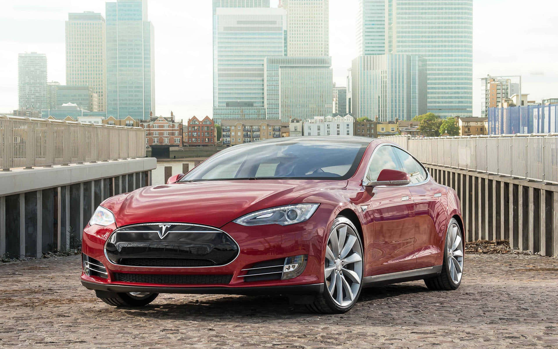 tesla 桌布 壁紙 — 一架亮麗的紅色Tesla Model S停泊在鵝卵石橋上，背後是現代摩天大樓。
