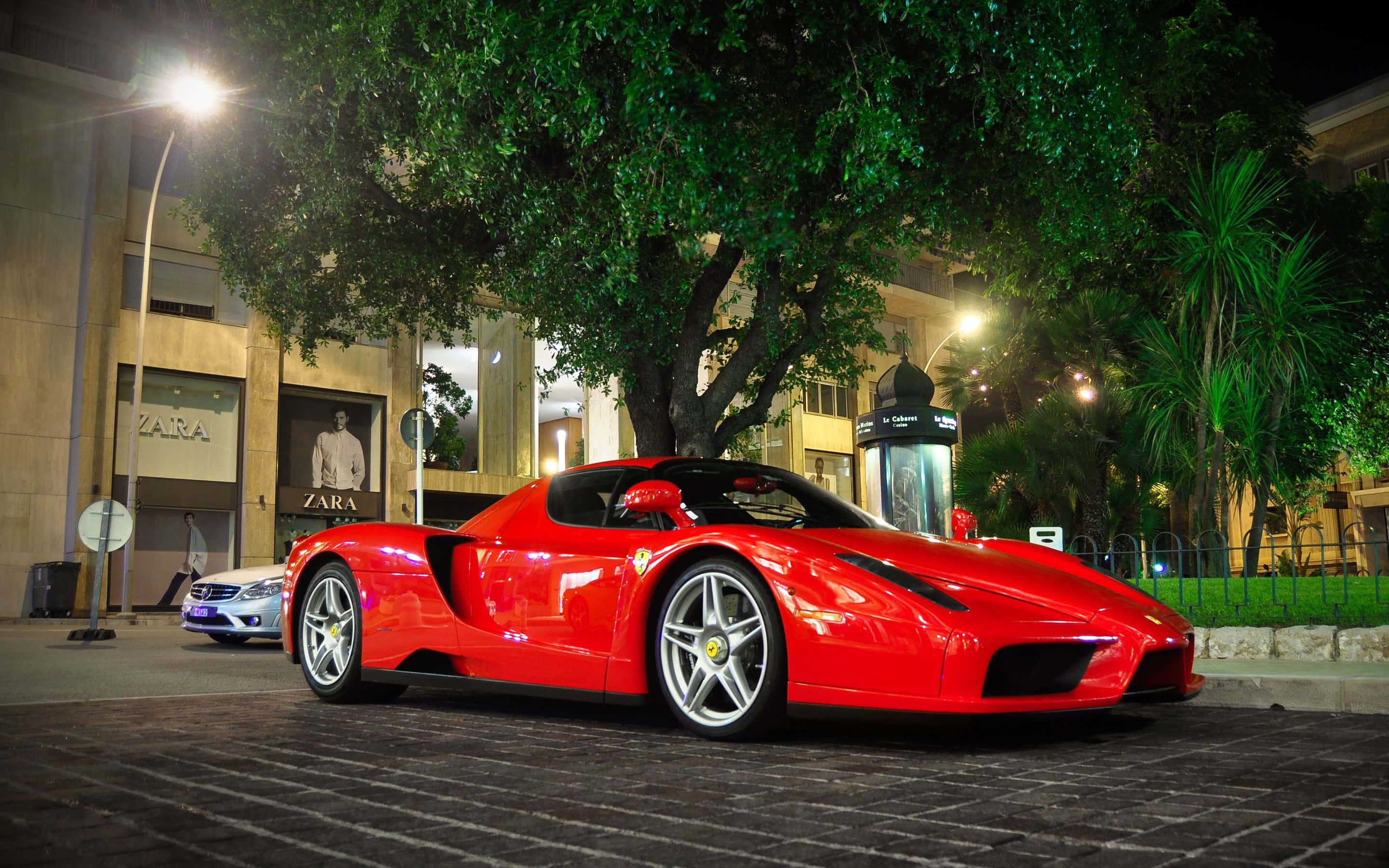 Ferrari Enzo 桌布 壁紙 — 一架亮麗的紅色法拉利Enzo在夜晚停泊在鵝卵石街道上，位於一棵大樹下，背景是燈火通明的商店門面。