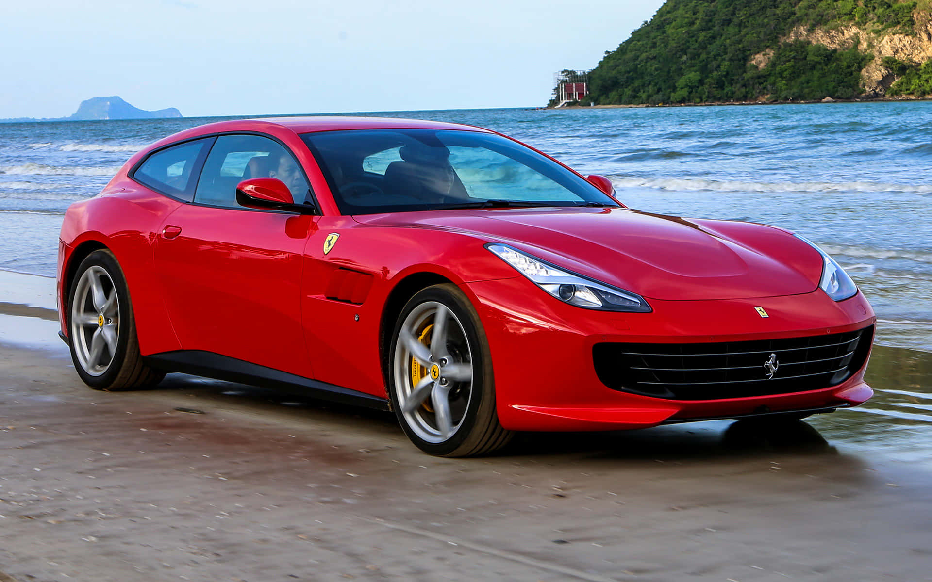 Ferrari Gtc4lusso 桌布 壁紙 — 一架亮麗的紅色法拉利跑車停泊在濕潤的沙灘上，背景有海浪和綠色山丘。