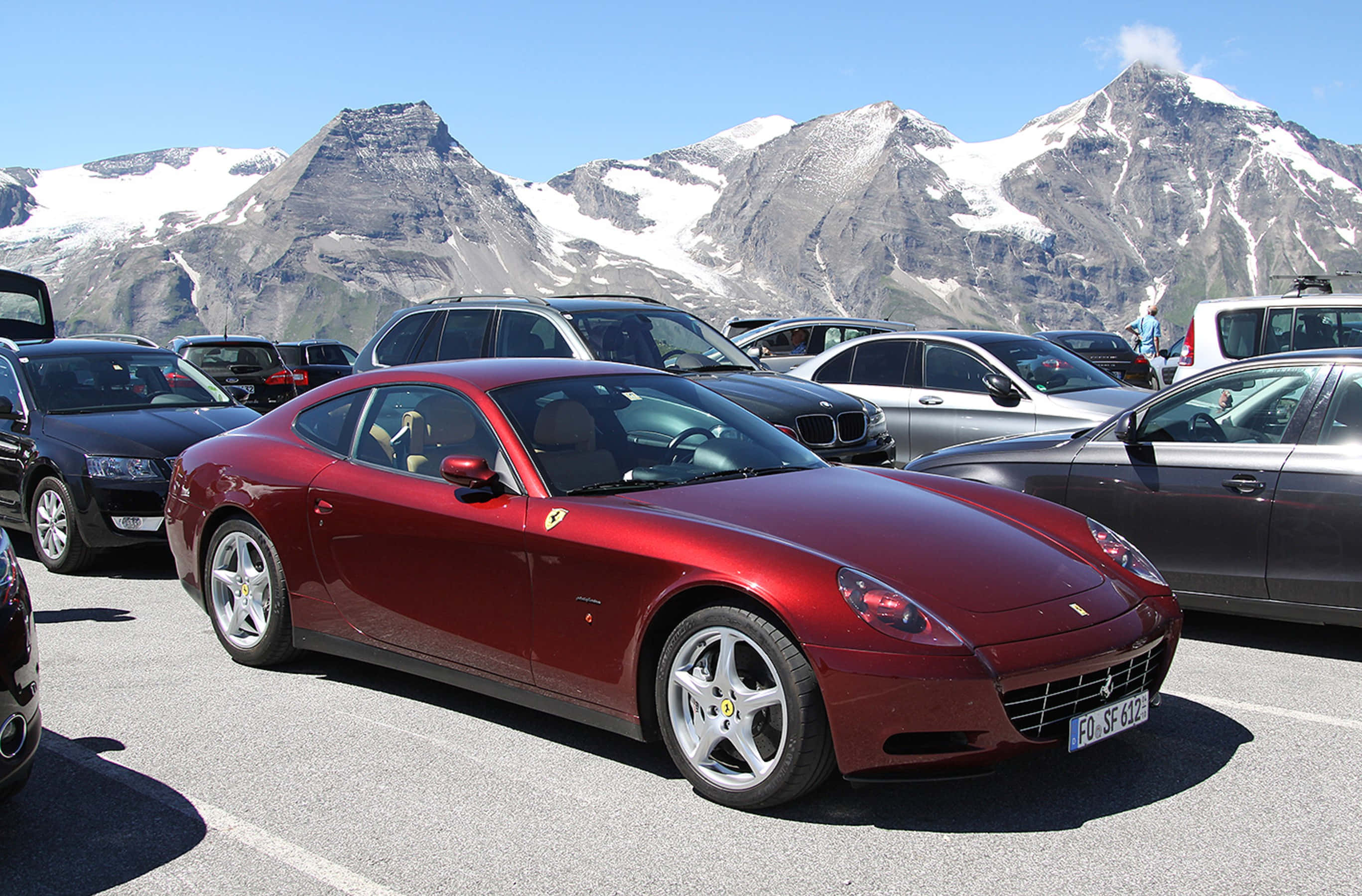 Ferrari 612 Scaglietti 桌布 壁紙 — 一架亮麗的紅色法拉利跑車停泊在阿爾卑斯山區的停車場，背景是白雪覆蓋的山峰和晴朗的藍天。