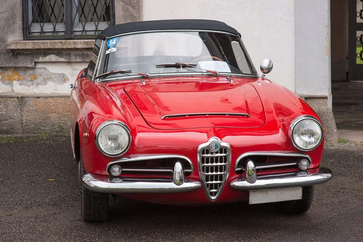 Alfa Romeo Spider 桌布 壁紙 — 一架亮麗的紅色經典愛快羅密歐Spider開篷車停泊在潮濕的街道上，旁邊是一棟風化的建築物。