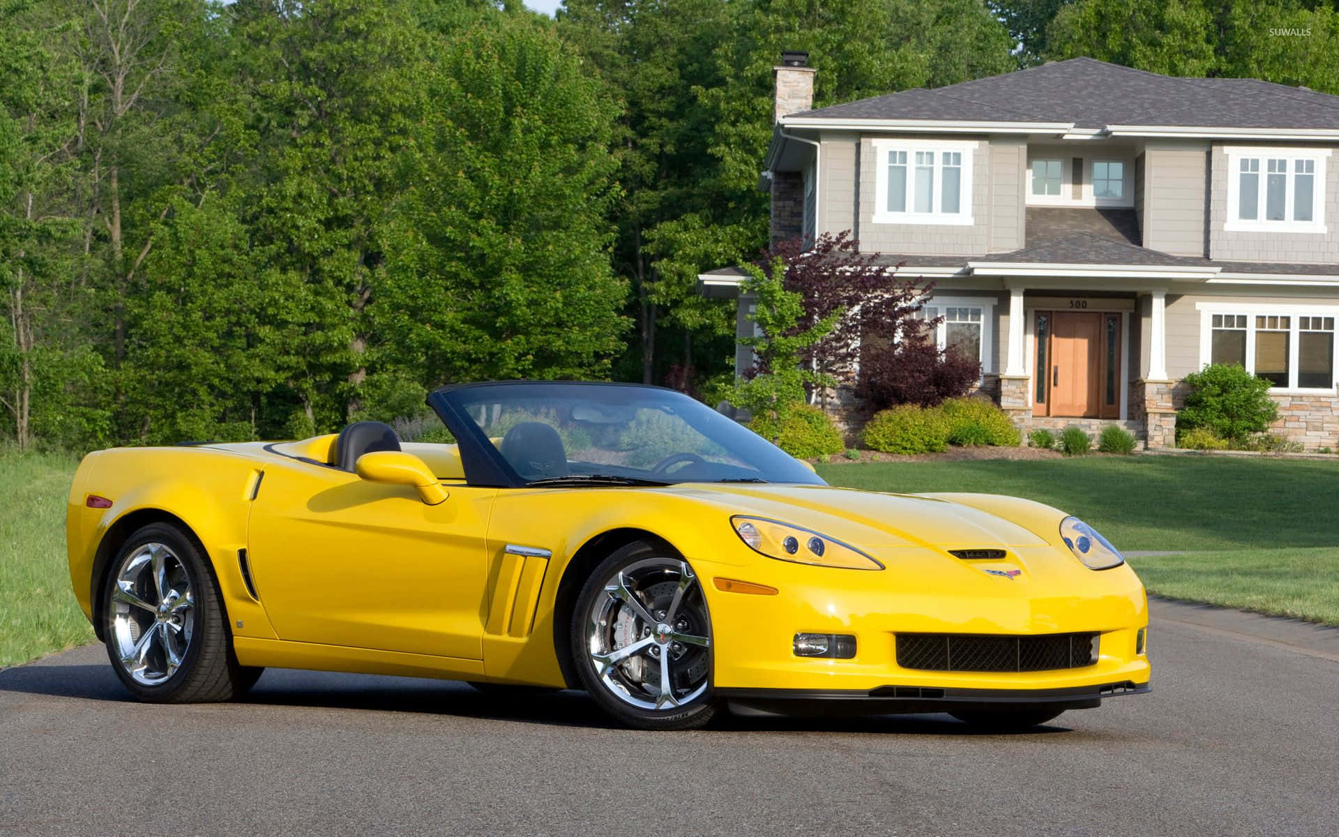 Chevrolet Corvette Grand Sport 桌布 壁紙 — 一架亮黃色的雪佛蘭克爾維特敞篷車停泊在郊區房屋前的車道上，周圍有茂密的綠樹。