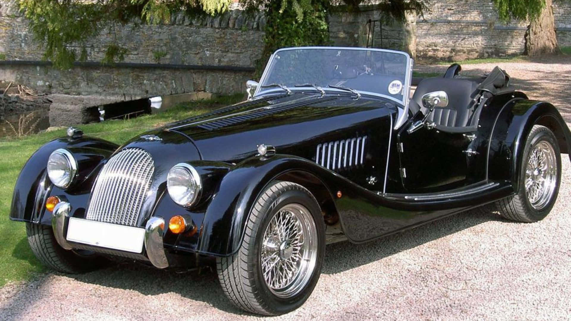 Morgan Roadster 桌布 壁紙 — 一架亮黑色經典摩根開篷跑車停泊在碎石路上，旁邊有石牆和綠草。