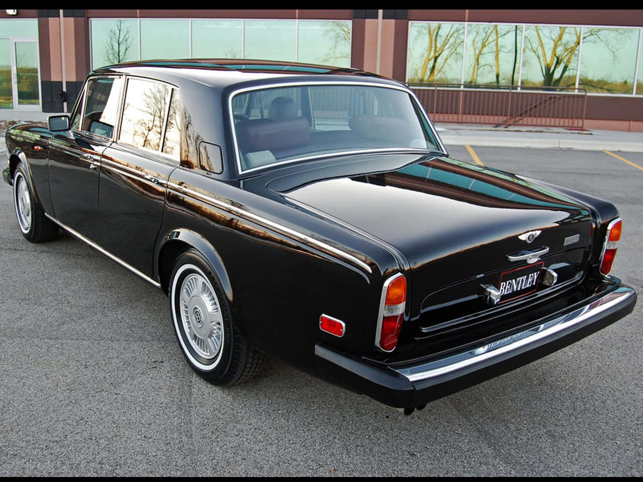 Bentley T2 桌布 壁紙 — 一架亮黑色經典賓利房車停泊在現代化停車場，背景有反光窗戶和光禿禿的樹木。