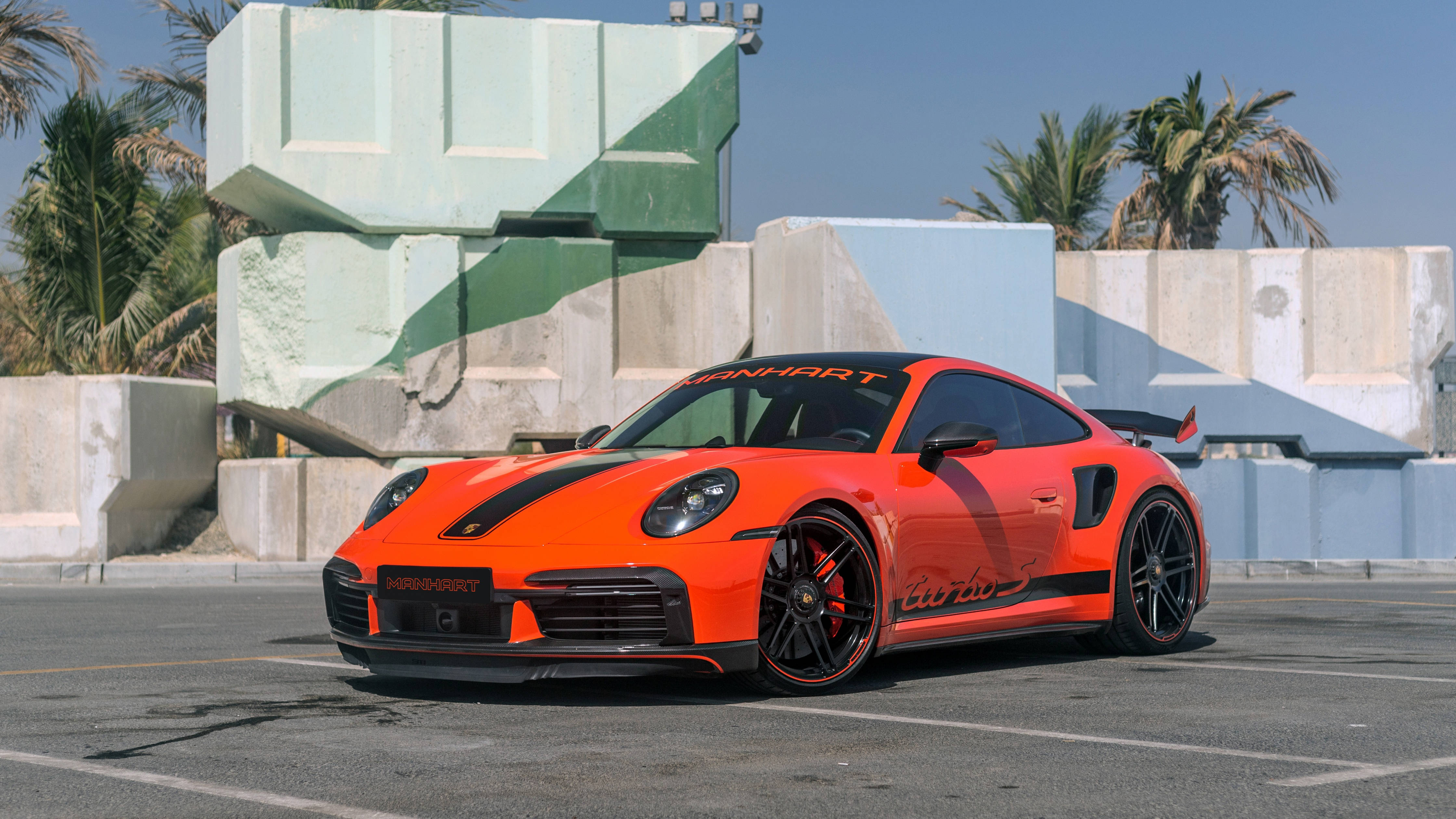 4K 桌布 壁紙 — 一架充滿活力的橙色Manhart改裝保時捷911 Turbo S停泊在陽光普照的混凝土停車場，背景有棕櫚樹和幾何結構。